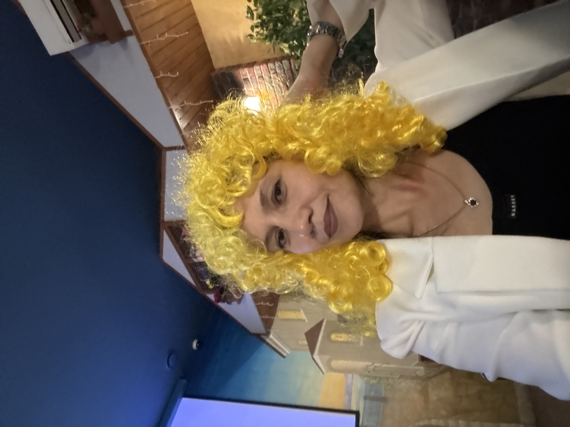 A woman with bright yellow curly hair and a white jacket is in a dimly lit indoor space. The background features a mural of buildings, possibly depicting a European street scene, and wooden paneling with decorative string lights. The woman is looking towards the camera and has her right hand on her head. There is a dark ceiling with some lighting fixtures visible. An air conditioning unit is mounted on the wall in the upper right corner.