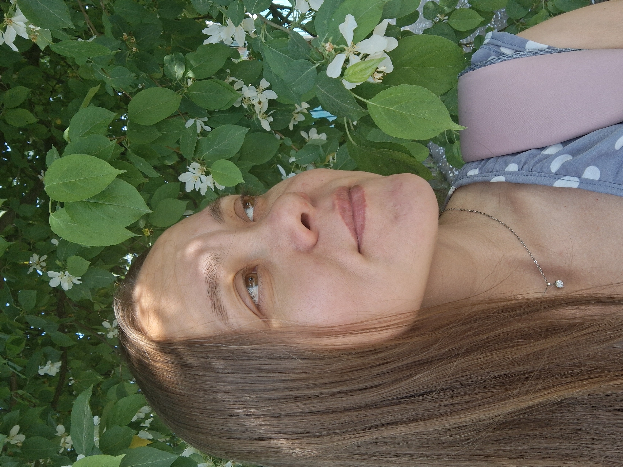 A medium shot depicts a woman lying on her side among a profusion of green leaves and white flowers. The background is a dense collection of vegetation. The woman has light skin, brown hair, and is wearing a gray and white polka-dot garment with a pink element. A delicate silver chain with a small stone pendant adorns her neck. The lighting is bright and appears to be natural daylight. The photo was taken in the city of Chaikovsky, Russia.