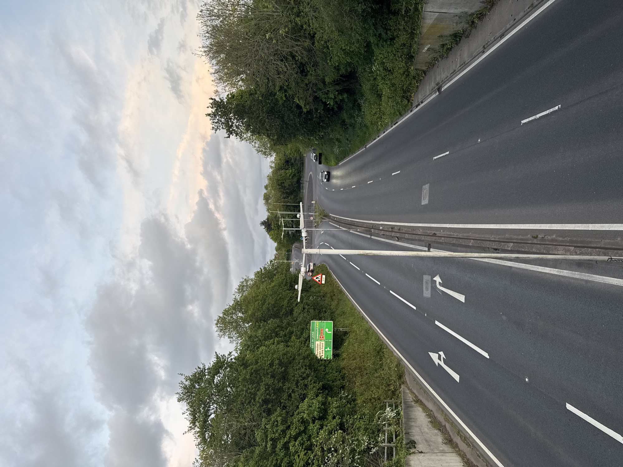 A vertical, eye-level, wide-angle shot captures a multi-lane highway winding through a lush, green forested area under a sky filled with a combination of dark and light clouds. The highway appears to be divided by a median, with white lane markings and directional arrows painted on the road surface. Signage is visible, including a green road sign with white lettering and symbols indicating directions to various locations such as Preston and other areas designated as "A47." The surrounding environment includes dense foliage, suggesting a rural or suburban location. Two cars are visible in the distance. The highway is flanked by concrete barriers. The location appears to be in the United Kingdom. Specific references to the city of Great Yarmouth could not be confirmed with certainty.