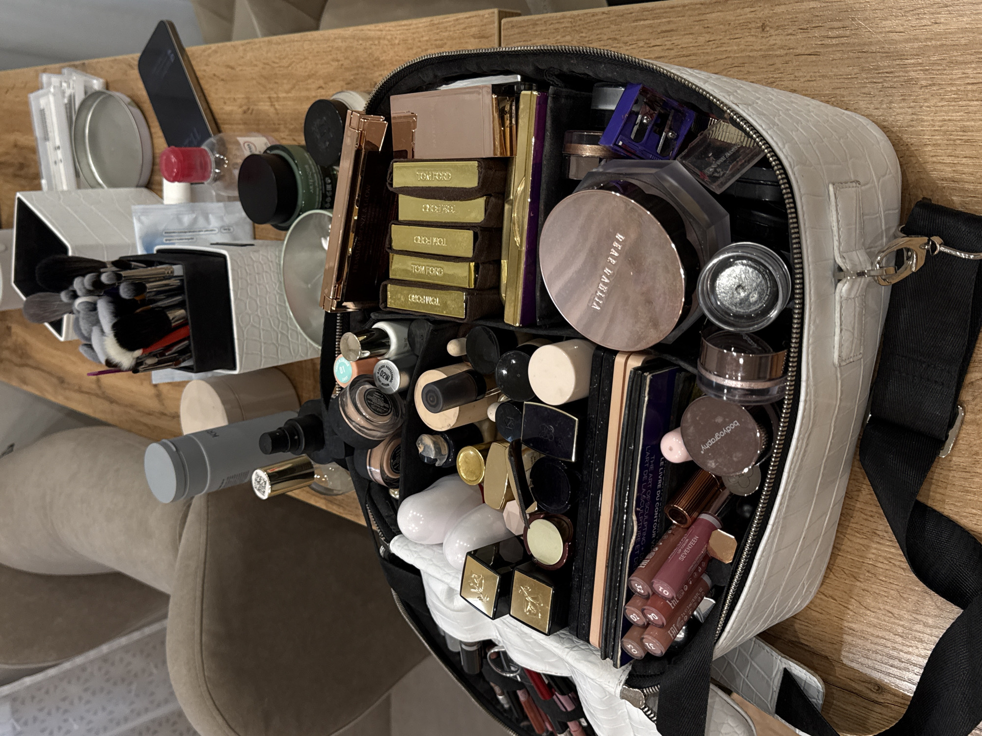 This is a high-angle, close-up shot of an open, white, crocodile-embossed makeup bag filled with various makeup products and brushes. The bag is resting on a light brown wooden surface, likely a table or counter. The lighting suggests it might be daytime, indoors.
The contents of the bag are abundant and varied, including several Tom Ford branded items, a large "DEAR NATALIA" labeled powder compact, lipsticks, various eyeshadow palettes, brushes, and other cosmetic containers. Some visible text includes "TOM FORD," "DEAR NATALIA," "bodyography," "SEVENTEEN," "LE LIVRE DU CONTOUR V," and "THE ART OF SCULPTING L'ART DE LA SCULPTURE ET." The overall impression is of a well-stocked makeup kit.