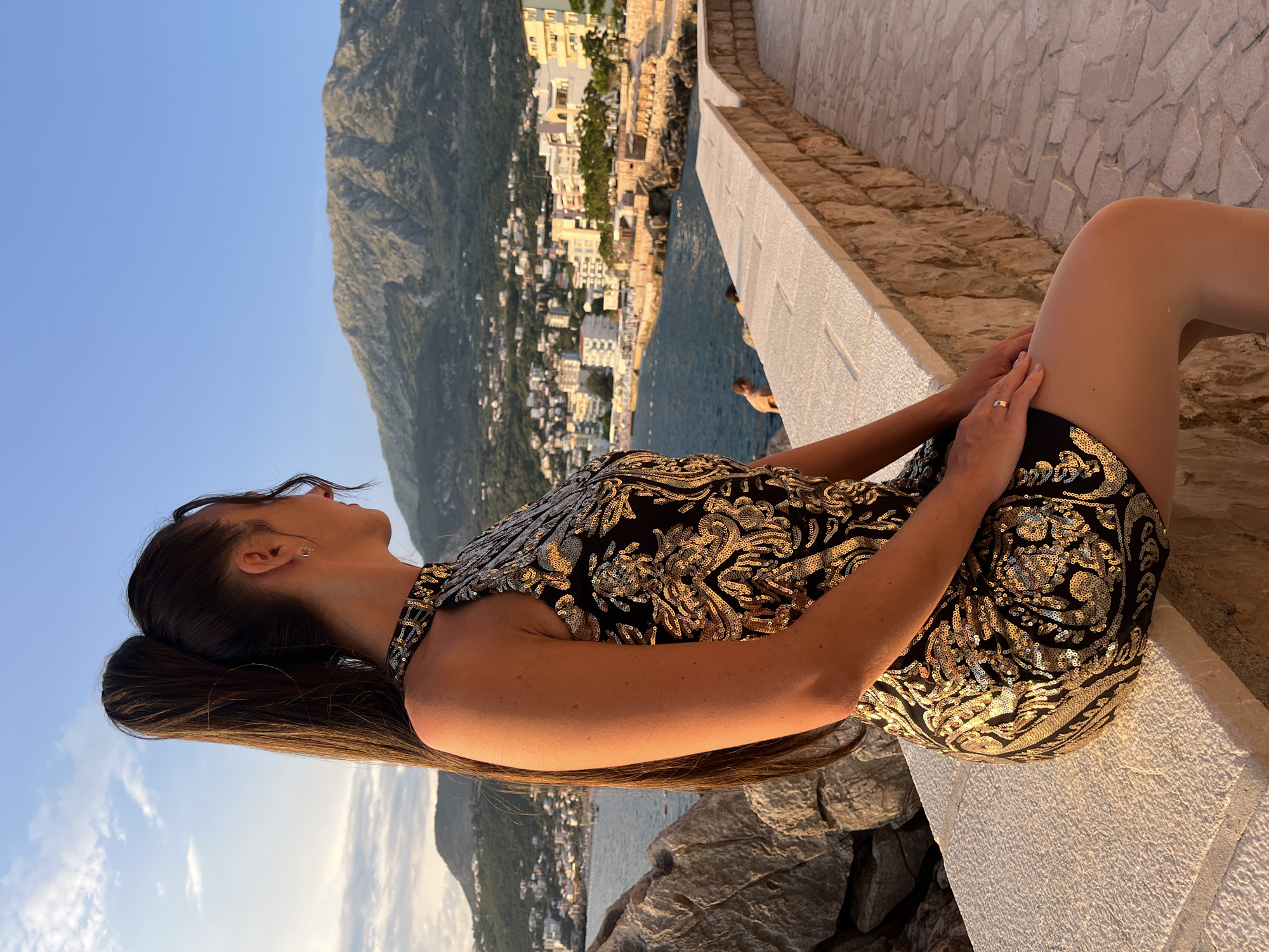 A woman with her back to the camera sits on a stone wall overlooking a coastal town. She has long, dark hair pulled into a high ponytail and wears a black sleeveless dress with a gold sequined pattern. Her left hand rests on her right leg, and she wears a gold ring on her left ring finger. The town features white and light-colored buildings clustered along the coastline, with a body of water in front of it. Behind the town, a large, rugged mountain dominates the landscape under a clear blue sky. The scene is illuminated by the warm light of what appears to be a sunset.
