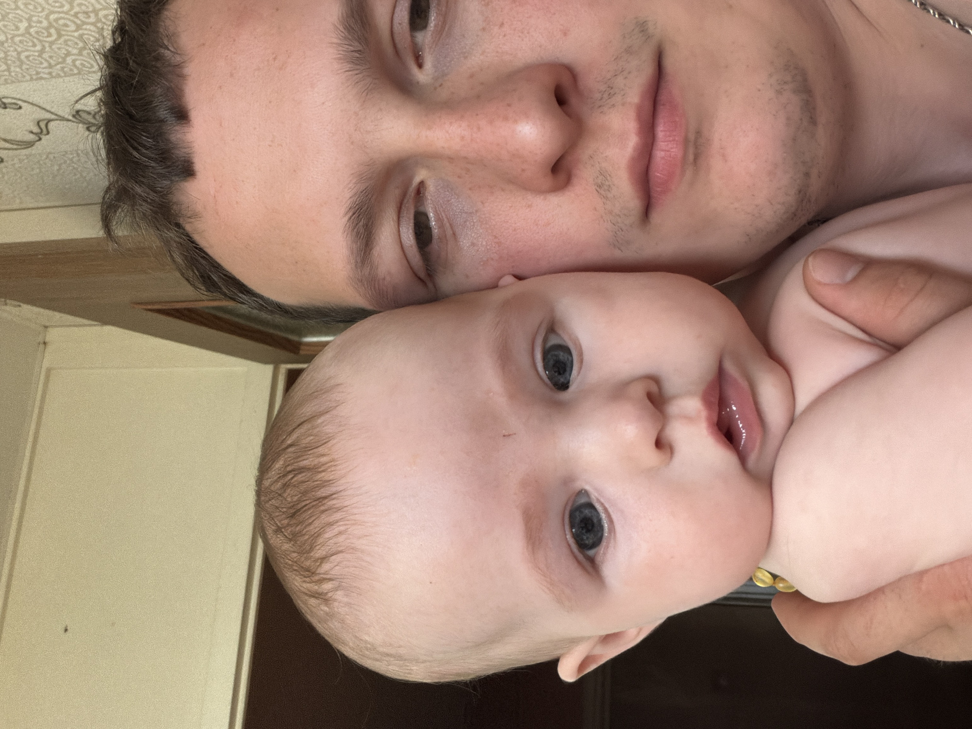 A close-up, head-and-shoulders selfie features a man and a baby. The man's face, partially obscured by the baby, occupies the right side of the frame. He has light skin, dark hair, and appears to be looking downwards. The baby, positioned on the left, is facing the camera with large, dark eyes and small lips. The background consists of a wall with a wood-paneled upper section and wallpapered lower section, typical of an interior residential setting in Chaikovsky, Russia.