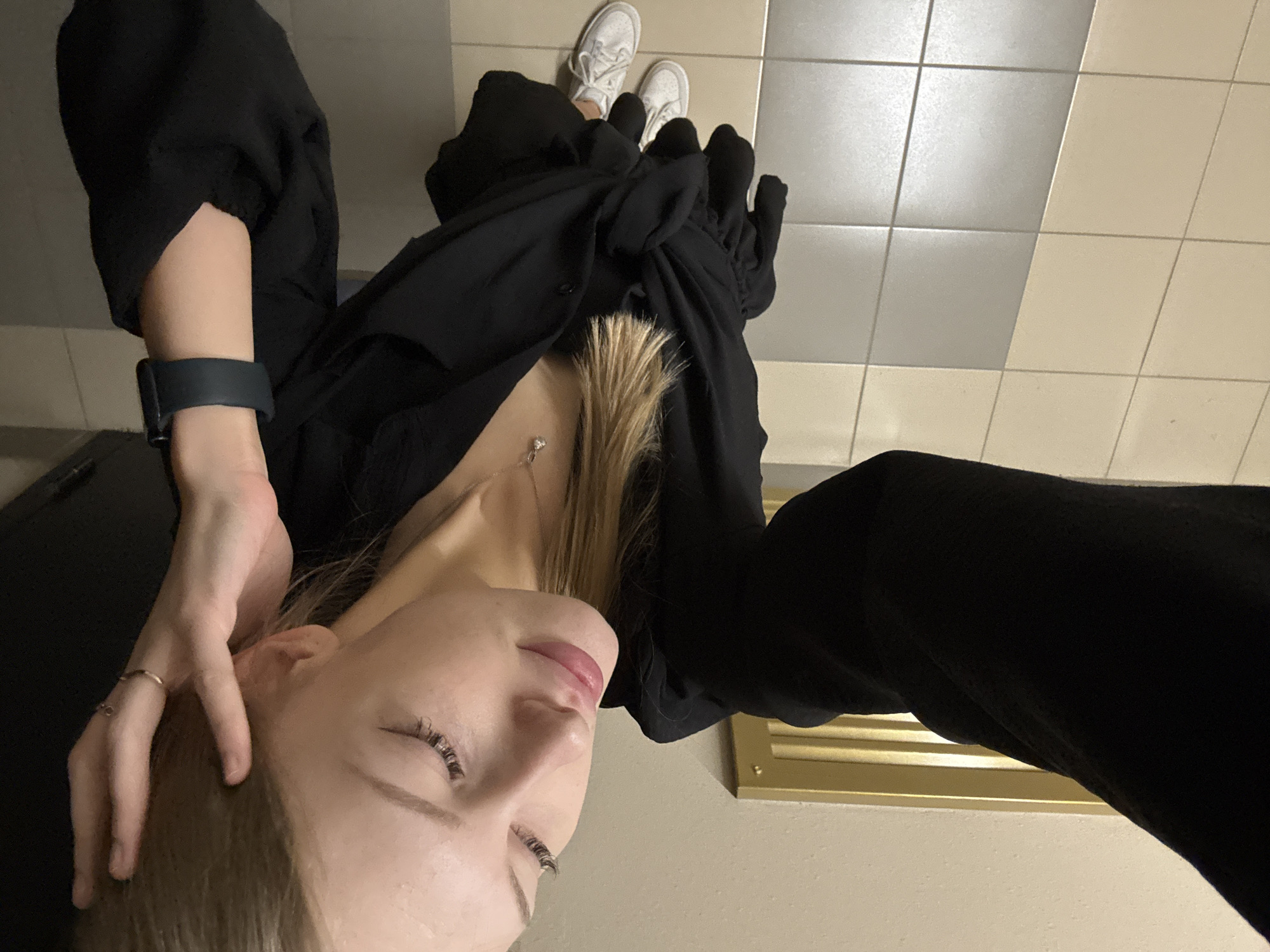 The image shows a young woman in a black outfit, lying down with her head tilted back. She has on white sneakers and a black watch on her left wrist. A delicate necklace with a small pendant rests on her chest. Her hand is casually placed on her head. The background appears to be an indoor tiled floor with squares of light and dark gray tiles. There is a golden vent or grate on the wall behind her. The lighting suggests it could be daytime indoors, or artificial lighting. The woman's eyes are closed, and her expression appears relaxed or perhaps even pensive. The overall mood is intimate and personal.