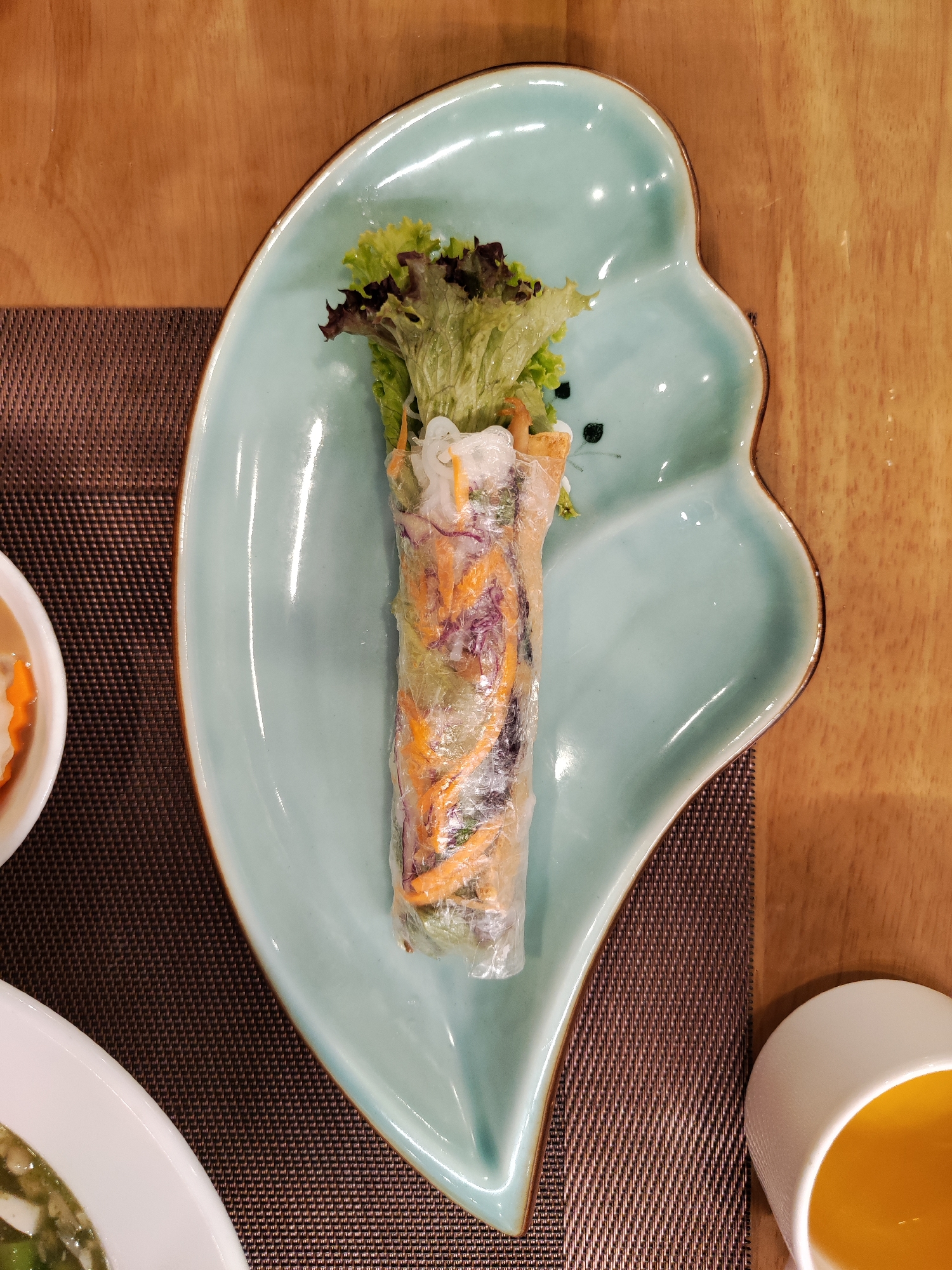A spring roll is presented on an ornate light-blue plate. The roll contains lettuce and orange and purple julienned vegetables, encased in translucent rice paper. The plate is positioned on a brown place mat, and it also has floral designs. A white bowl with a clear orange liquid and another white dish containing a green soup or sauce are partially visible to the lower left and right, respectively. The background consists of a wooden surface, likely a table. The scene appears to be set in a restaurant. The location is Da Lat, Vietnam.