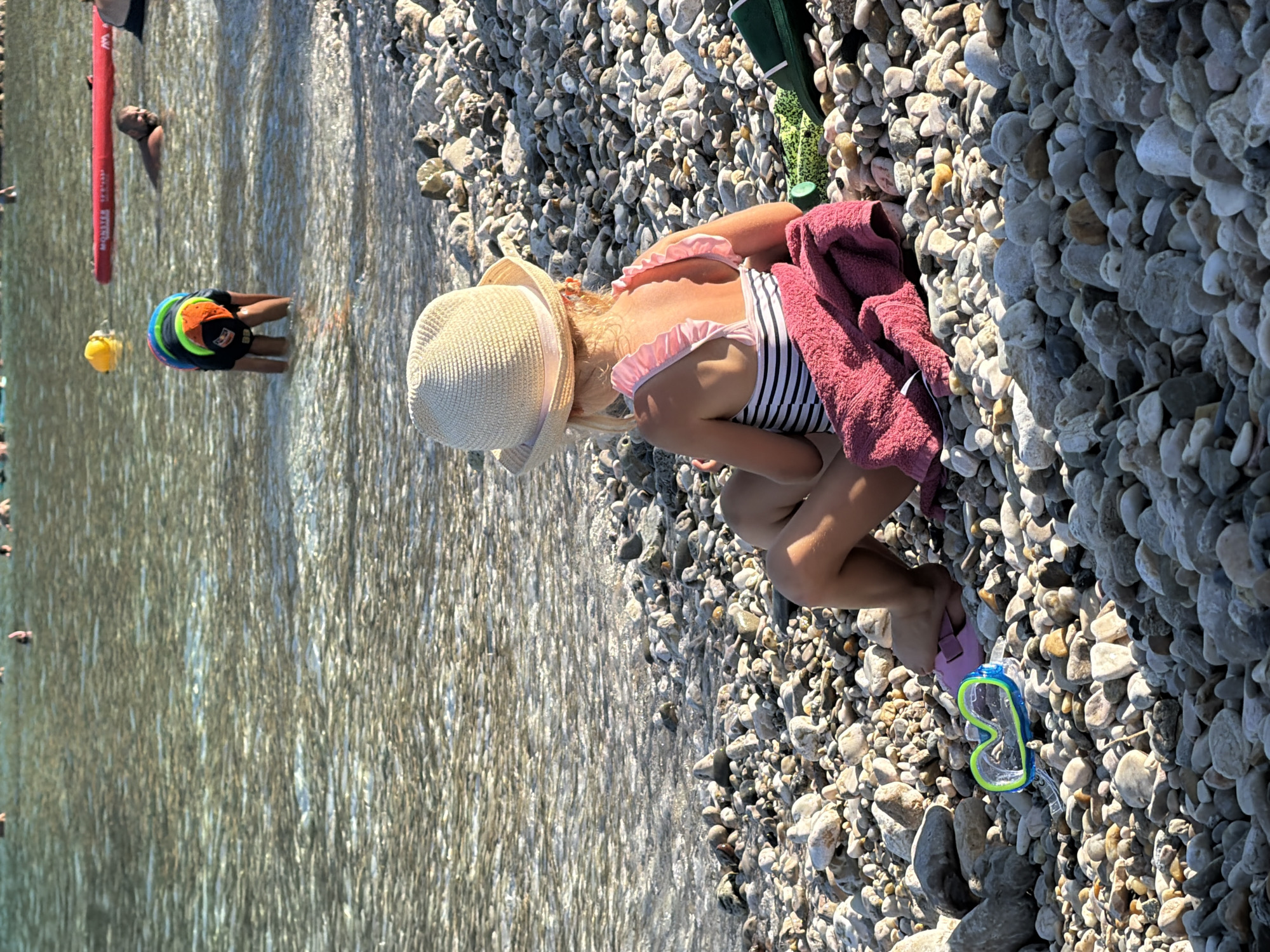 In this horizontal, eye-level shot, a young girl is crouched on a sun-drenched, pebble-strewn beach by the water's edge. She wears a striped swimsuit and a wide-brimmed straw hat adorned with a pink ribbon. A pink towel is draped over her shoulder. Her knees are bent, and her arms rest on the pebbles. Next to her, a pair of blue and green swim goggles lie on the stones.
In the shallow, clear water to the left of the frame, another child is visible wearing a life vest with multiple colorful flotation rings. Further left, a person is partially submerged in the water, their head and shoulders visible. A red inflatable object, possibly a paddleboard or pool float, is also in the water. The scene suggests a warm, sunny day, likely mid-afternoon given the strong shadows. The pebble beach and clear water are characteristic of coastal areas in Croatia, consistent with the Općina Baška location. The overall atmosphere appears relaxed and indicative of a leisurely day at the beach. There is no visible text in the image.