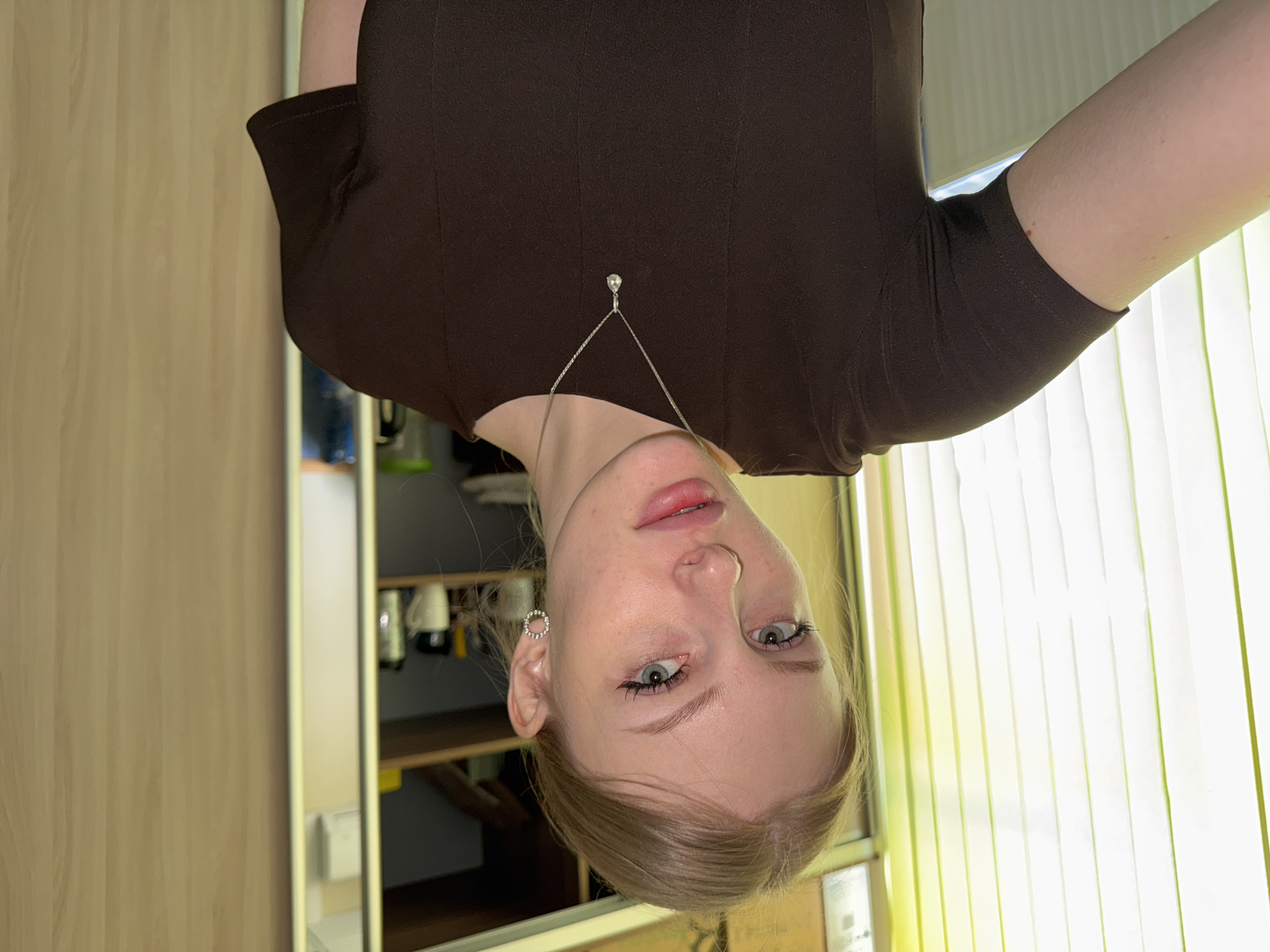 A fair-skinned woman with blonde hair is seen in a selfie-style photograph. She is wearing a brown top and a delicate necklace with a small pendant. Her makeup is minimal, and she has an earring in her left ear. The background includes what appears to be a cabinet or closet with items on shelves, and a window with vertical blinds. The location appears to be inside a building, possibly a residence. There is no definitive evidence to suggest this photo was taken in Perm, Russia.