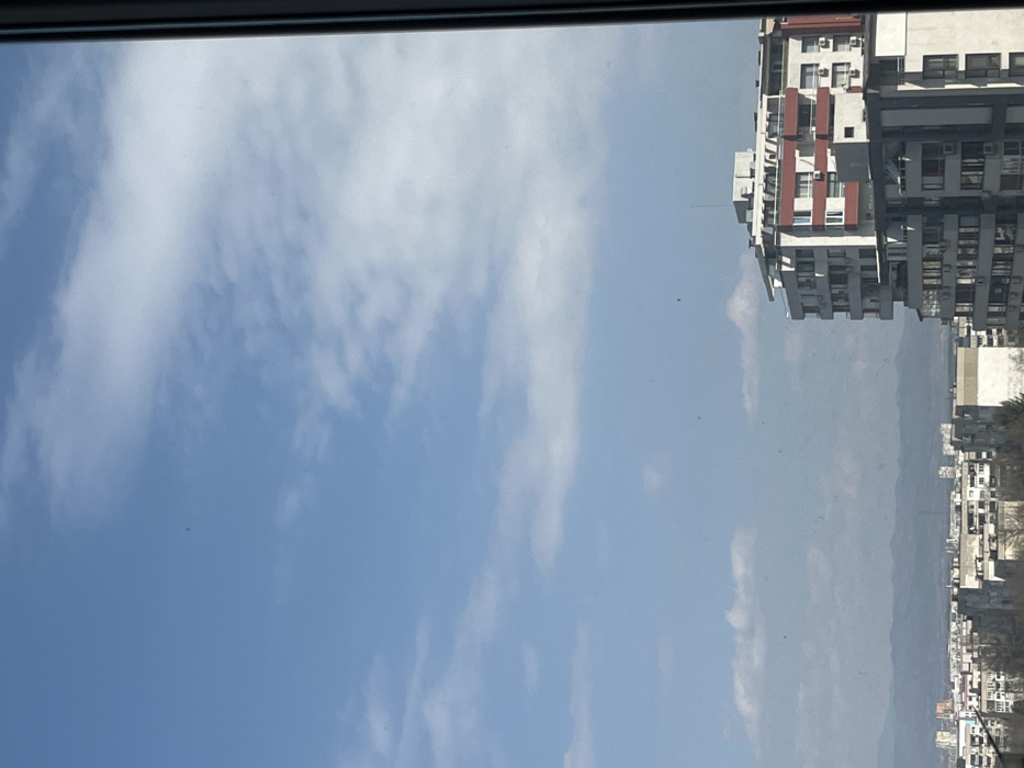 The image displays an urban cityscape under a partly cloudy blue sky, viewed from a high vantage point, likely through a window with a dark vertical frame visible on the right edge. Numerous small, indistinct specks are present across the image, consistent with dust or debris on a window pane.
In the midground, the city of Sofia, Bulgaria, stretches across the frame. A prominent multi-story residential building occupies the right portion of the image. This building features white facades, several floors with balconies, and distinctive red-colored panels or balcony railings on its upper sections. Numerous air conditioning units are visible on its exterior walls. Below and to the left of this building, a dense cluster of other multi-story residential and commercial buildings, predominantly in shades of white, grey, and beige, extends towards the horizon. Bare tree branches are discernible among these structures.
In the background, a distant, hazy mountain range, characteristic of the Vitosha mountains, forms the horizon line under the expansive sky. No individuals, explicit actions, or interactions are visible within the scene. The lighting indicates clear daylight conditions.