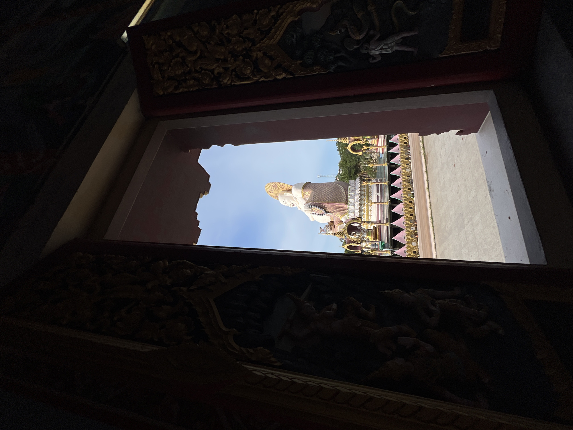 The image shows a view through an ornate, dark wooden doorway, looking out onto a large, white, seated Buddha statue. The Buddha is adorned with a pink and gold robe and a golden halo. It is situated on a platform surrounded by colorful decorations and smaller ornate structures. The background features a clear blue sky and some greenery. The foreground is dominated by the dark, carved interior of the doorway, with the sunlight creating a stark contrast with the illuminated Buddha and exterior. This scene is located at the Big Buddha Temple in Baan Plai Laem, Thailand.