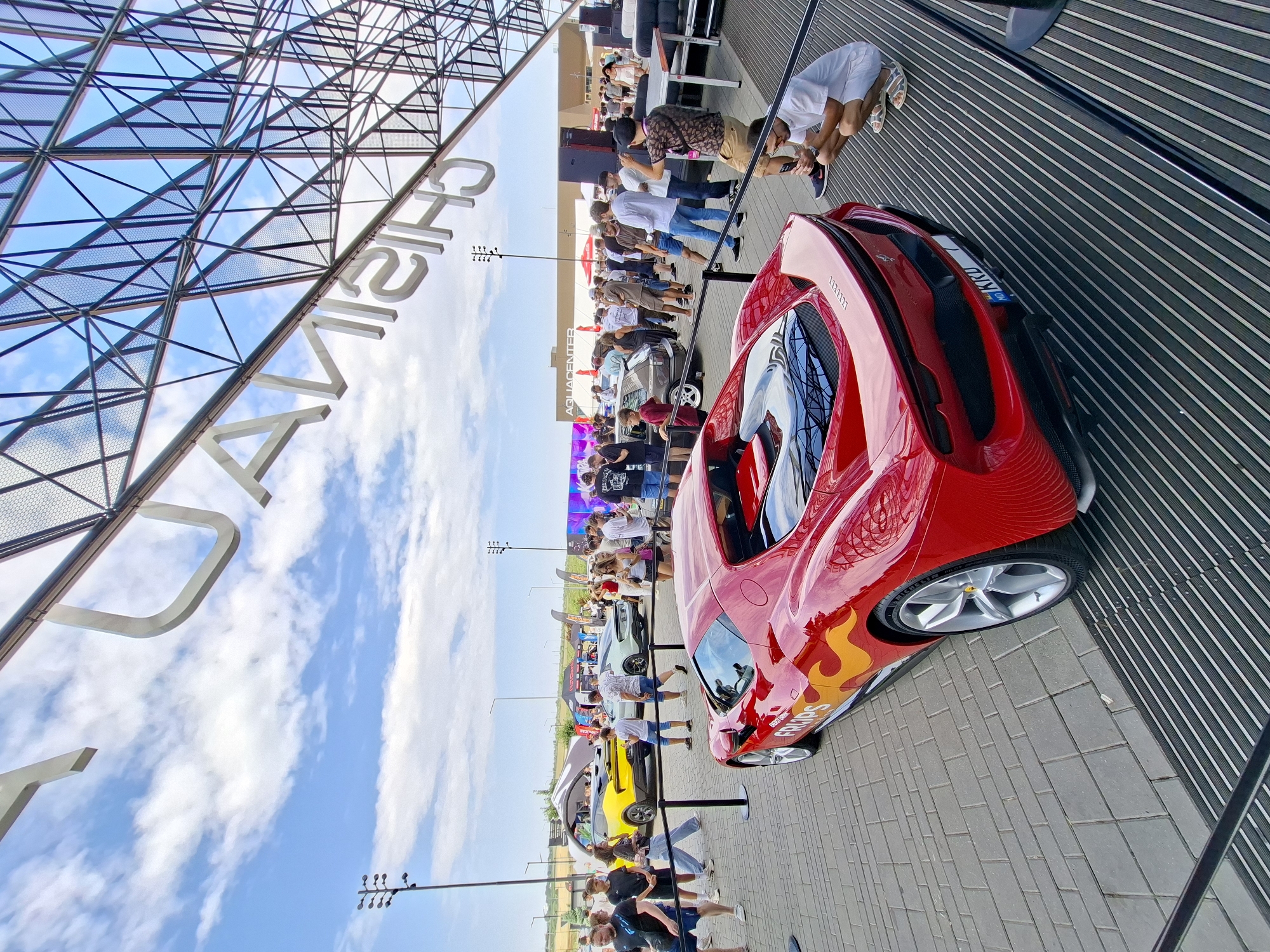 A red Ferrari sports car with flame decals on its side is parked on a brick pathway. The car has a license plate that reads "MD KRD". In the background, a large crowd of people are gathered, and several other cars are on display. There is a building with a large sign that says "AQUACENTER". The scene appears to be an outdoor event or car show. The sky is blue with scattered clouds. The event is taking place in the city of Stăuceni, Moldova. A man is crouching down in the foreground, appearing to take a picture of the car with his phone.