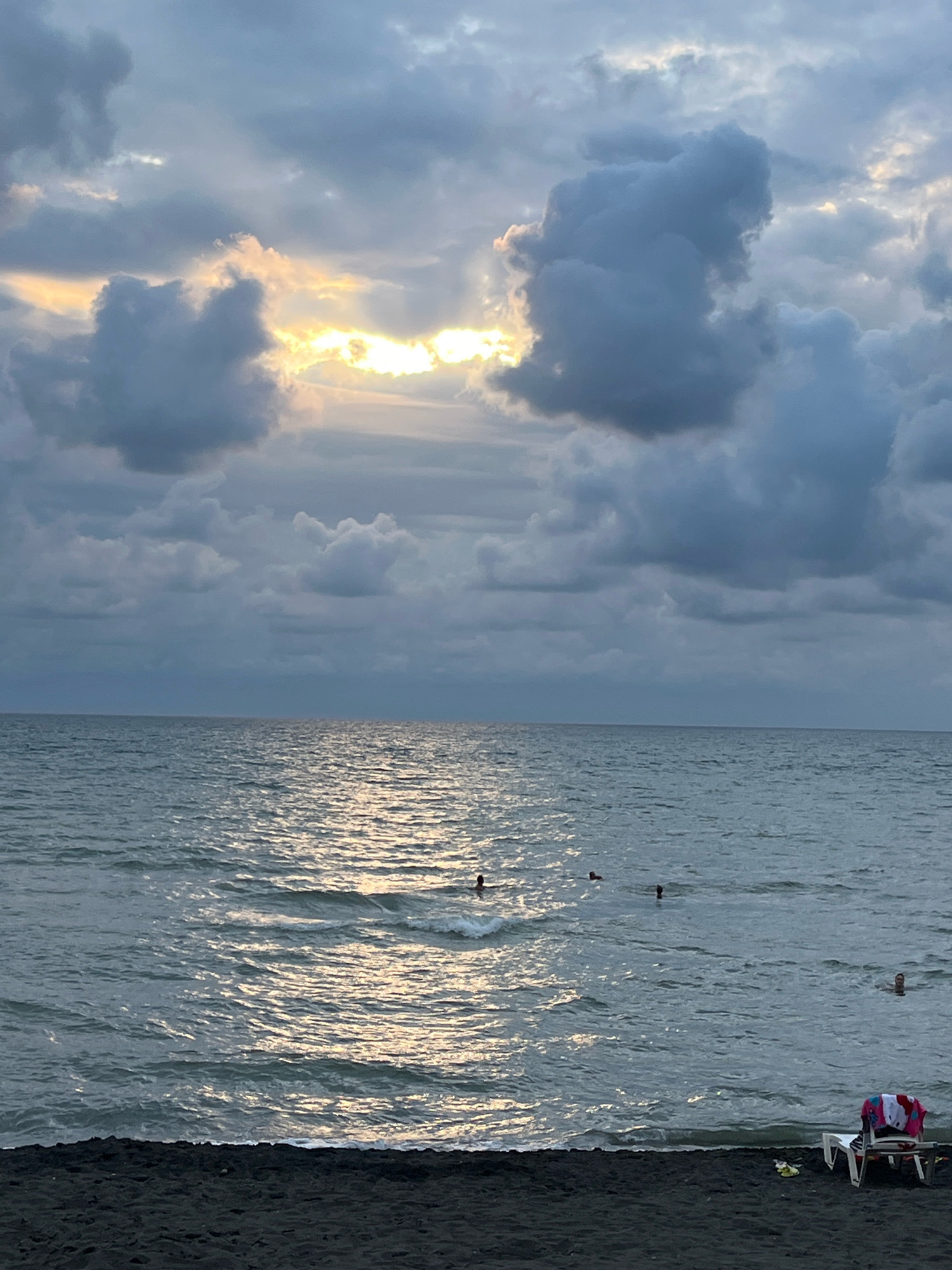 A dark, sandy beach in Ureki, Georgia, meets the calm, shimmering waters of the Black Sea under a cloudy, overcast sky. Patches of sunlight break through the dense clouds, casting a warm glow on the sea's surface. Several people are swimming in the water, their bodies silhouetted against the light. On the beach, a white lounge chair is set up with towels and a bag. The scene is peaceful, with the gentle waves and diffused sunlight creating a serene atmosphere.