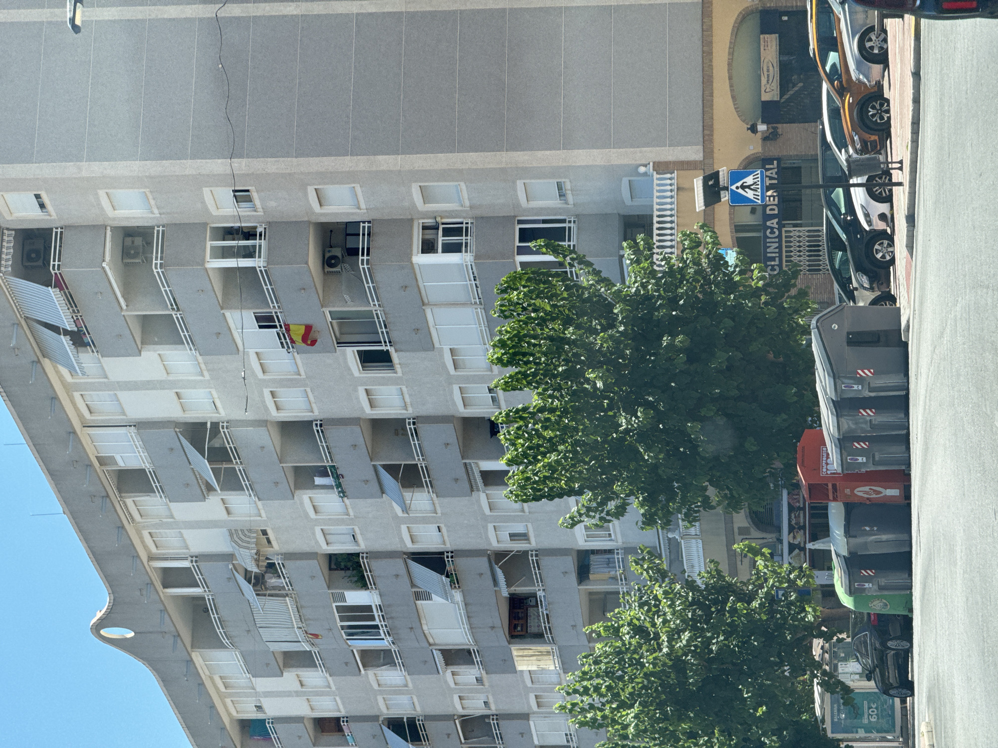 The image depicts a residential building in a Spanish town, likely La Vila Joiosa, under a clear blue sky. The building is tall and gray, with numerous balconies and windows. Some balconies have awnings, and a Spanish flag is visible hanging from one of them. Lush green trees are in the foreground, obscuring parts of the building.
On the street in front of the building, several cars are parked. A pedestrian crossing sign and a sign for "CLINICA DENTAL" are clearly visible. The presence of multiple large trash bins suggests a public urban setting. The sunlight indicates it is daytime, and the bright sky suggests fair weather. The scene appears to be a typical street view in a Spanish town, likely during the day, with no obvious signs of any specific event or activity beyond daily life. The overall mood is neutral, conveying an everyday urban environment.
Visible text includes "CLINICA DENTAL," a pedestrian crossing sign, and text on the trash bins and a sign on the street that reads "BALEARIA" and "60c".