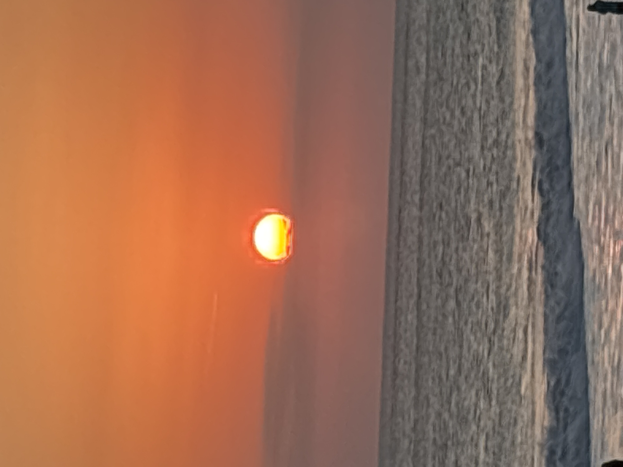 The image depicts a sunset over the ocean in North Kuta, Indonesia. The sun, a bright orange orb, is partially obscured by a thin layer of cloud and is low on the horizon. The sky is a gradient of orange and pink. The ocean is a muted grey with white waves breaking on the shore. Two figures are visible on the beach in the foreground, silhouetted against the fading light. One figure is standing on the left, and another is standing on the right. The scene is serene and peaceful, capturing the beauty of a tropical sunset.