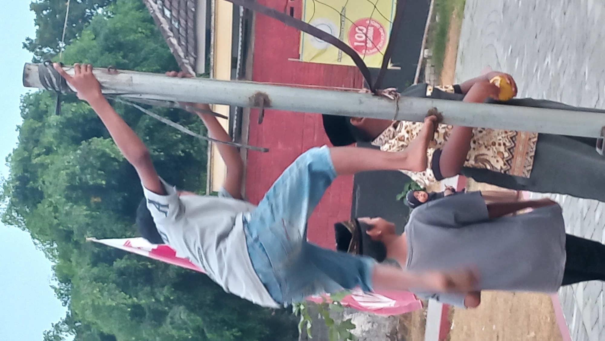 In the city of Pare, Indonesia, a young boy is captured mid-climb up a metal pole, his bare feet gripping the rough surface. He is wearing a light grey t-shirt and light blue denim shorts, with a black skullcap perched on his head. His left arm is extended, grasping the pole for support, while his right arm is bent, also holding on. To the left of the boy, a red and white flag is visible, partially obscured by trees. In the background, a brick building with a tiled roof stands, and a yellow banner with red text is displayed on a net. Another young boy, wearing a grey t-shirt and black pants, stands facing away from the camera. A third individual, seen from the chest up, wears a patterned shirt and a black skullcap, holding a yellow object in their hand. The ground beneath them is paved with interlocking bricks, and sparse green foliage is scattered around. The sky above is overcast with a pale blue hue.