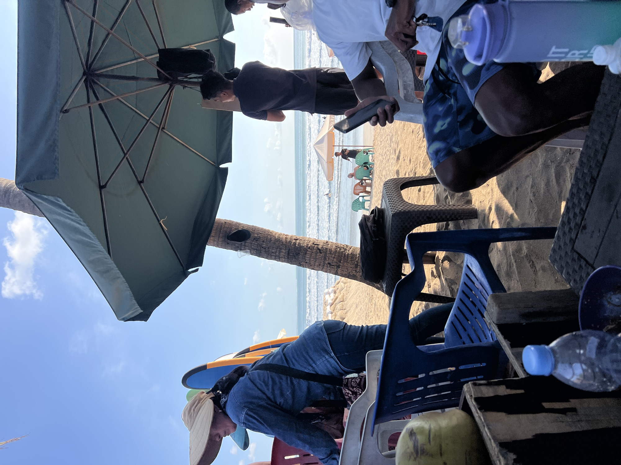 This is a photograph taken from a low angle on a sandy beach, likely in Kuta, Indonesia, with the ocean visible in the background. The image is dominated by a large, dark green umbrella providing shade. Several people are present in the scene, some sitting and others standing.
In the foreground, a person wearing a wide-brimmed hat and a denim jacket is partially visible, leaning over a blue plastic chair. Next to them, a coconut sits on a wooden crate. Another blue plastic chair is in view, and further to the right, two people are seated. One is wearing patterned swim shorts and holding a smartphone, while another person in a white shirt is partially visible. A light blue water bottle is also prominent in the foreground.
In the middle ground, under the umbrella, several more chairs and tables are arranged on the sand. A palm tree trunk rises near the umbrella. Further back, on the beach, a group of people are seated at a small table under a tan umbrella. In the distance, the ocean stretches out with gentle waves, and a few individuals are in the water. Surfboards are visible near the seated person in the denim jacket. The sky above is a clear blue with some scattered clouds.