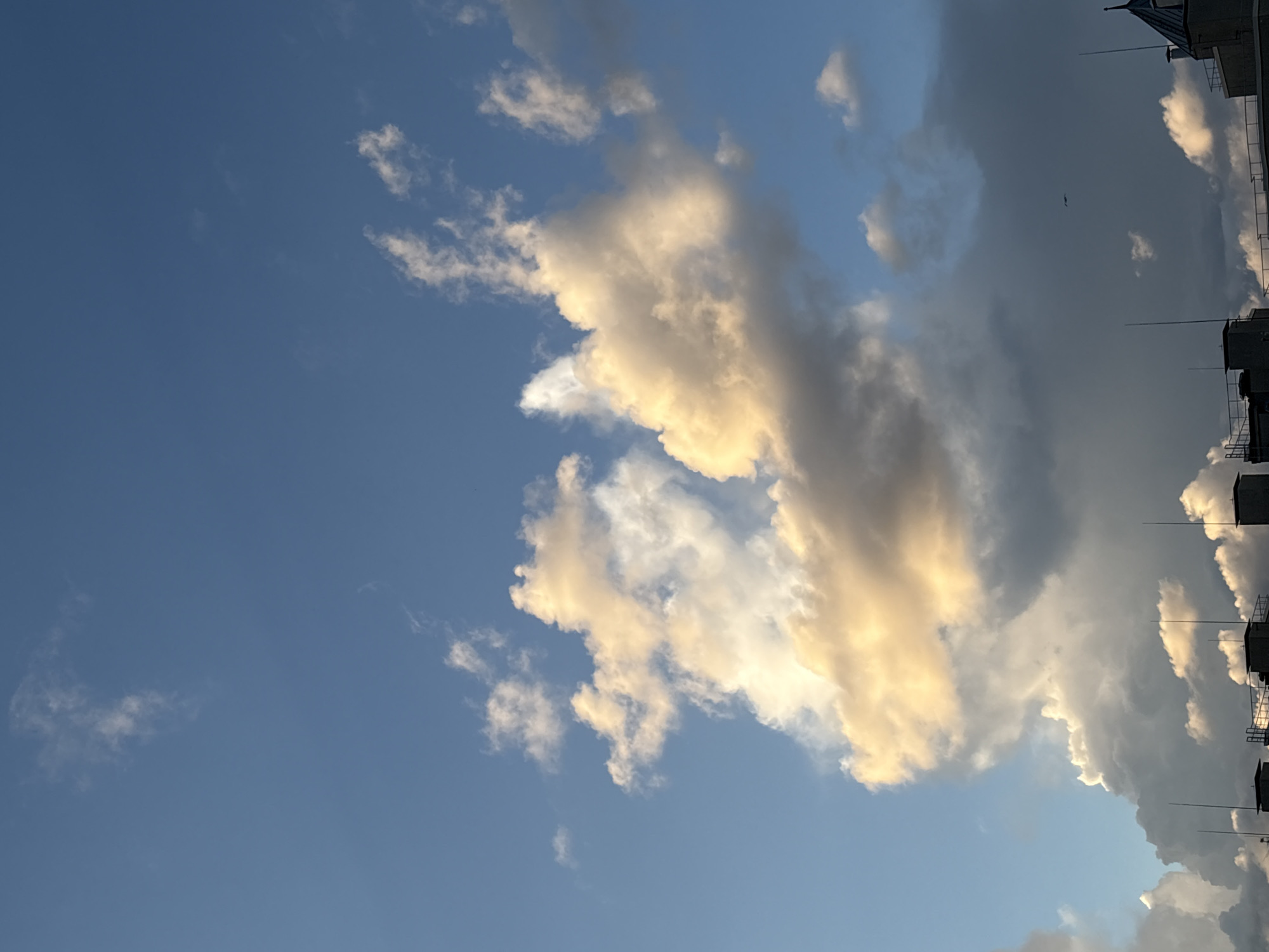 A photograph of the sky above Saint Petersburg, Russia, is depicted. The image features a vast expanse of blue sky partially obscured by a dramatic arrangement of clouds. A large cluster of clouds dominates the center of the frame, with the sun illuminating their edges, casting a warm, golden-yellow glow. These clouds appear to be illuminated from the side or back, suggesting it is either sunrise or sunset. Below these, darker, more ominous clouds are visible, indicating a change in weather. The bottom of the image reveals the rooftops of buildings in Saint Petersburg, characterized by their flat structures and prominent antennae. The overall scene captures a moment of transition in the sky, with the interplay of light and shadow on the clouds creating a sense of depth and drama above the urban landscape.
