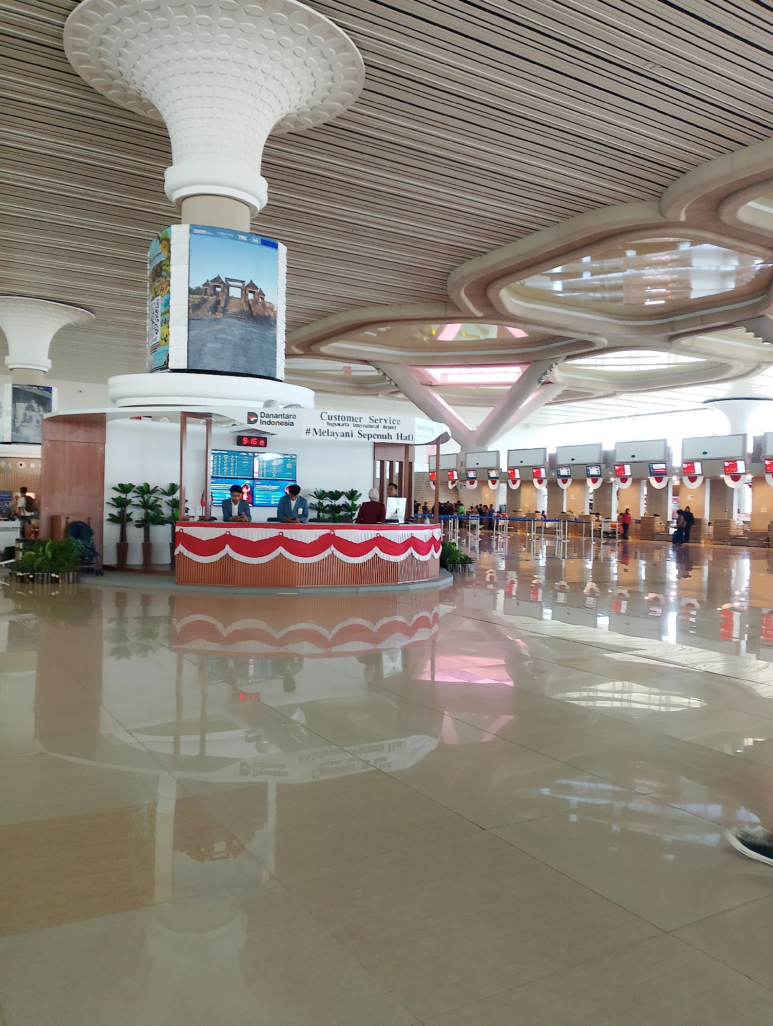 The image shows the interior of an airport terminal in Palihan, Indonesia. The scene is dominated by a polished, reflective floor that mirrors the ceiling architecture and the surrounding elements. In the foreground, a customer service counter is visible, decorated with red and white fabric. Two individuals in blue uniforms are attending the counter, and several potted plants are placed around it.
Behind the counter, a large digital display shows flight information. A prominent pillar in the center of the frame features a large cylindrical advertisement with an image of a historical building and text in Indonesian, including "Danantara Indonesia" and "#Melayani Sepenuh Hati". The ceiling is an elaborate structure with horizontal wooden slats and decorative, curved segments.
In the background, numerous check-in counters with digital screens are arranged in rows. Several passengers and staff members are present, suggesting a functional airport environment. The overall atmosphere appears bright and modern. The reflection on the floor indicates a well-maintained and spacious terminal.