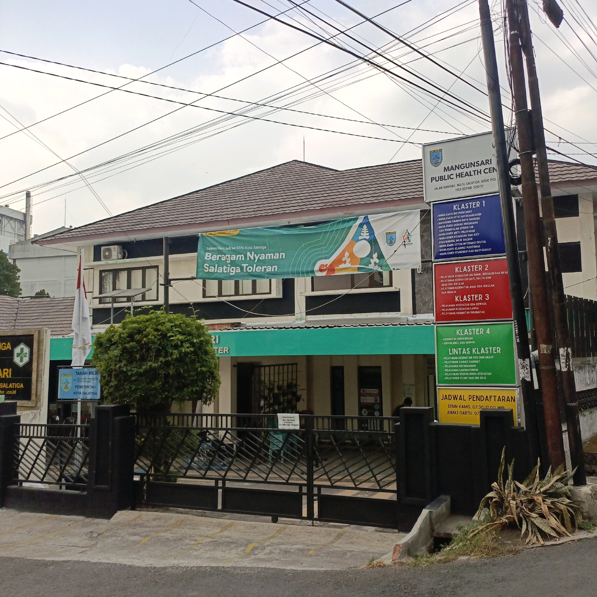 The image shows the exterior of the Mangunsari Public Health Center in Salatiga, Indonesia. The building is two stories tall with a dark tiled roof and a dark-colored facade. A large green banner is hung across the front of the building, displaying text that reads "Selamat Hari Jadi ke-1276 Kota Salatiga" and "Beragam Nyaman Salatiga Toleran." Multiple signs are posted on poles in front of the building, one of which identifies it as the "MANGUNSARI PUBLIC HEALTH CENTER" with an address and phone number. Other signs list "Klaster 1," "Klaster 2," "Klaster 3," "Klaster 4," and "Lintas Klaster," with bulleted lists of services under each. A yellow sign indicates "JADWAL PENDAFTARAN" (Registration Schedule). A white flag with red and white stripes is visible on a flagpole. In the foreground, a black metal fence encloses the property, and a paved area with yellow lines marking parking spots is visible. A small, overgrown plant with long, spiky leaves is situated next to a concrete curb. Power lines are prominent in the sky above the building. The scene appears to be captured during the daytime under an overcast sky.
