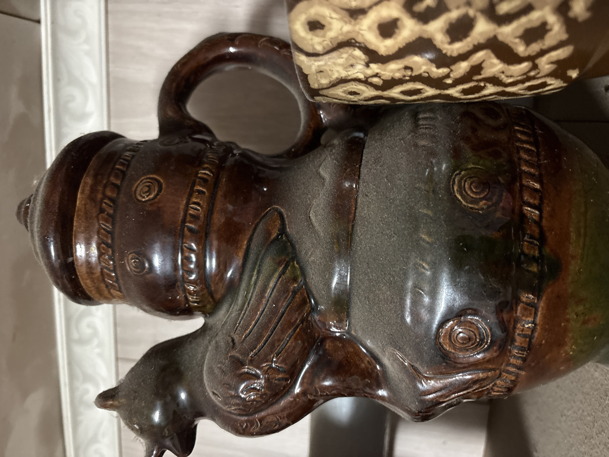 A close-up, eye-level shot shows a dusty, brown ceramic pitcher shaped like a bird, with intricate scrollwork and ribbed detailing. The pitcher has a long beak, feathered wings, and a lid with a rounded top. To the right of the bird pitcher, partially visible, is another brown ceramic vessel with a geometric pattern of light brown diamond shapes. The background consists of a light-colored, textured wall with a decorative white trim along the top. The scene appears to be indoors, possibly on a shelf or table. The pitcher's dusty surface suggests it has been in place for some time. The overall lighting is dim, with light reflecting off the glazed surfaces of the pottery. The location is identified as Ветлужский, Russia, suggesting the pitcher is an artifact from this region. There are no people or actions visible in the image.
