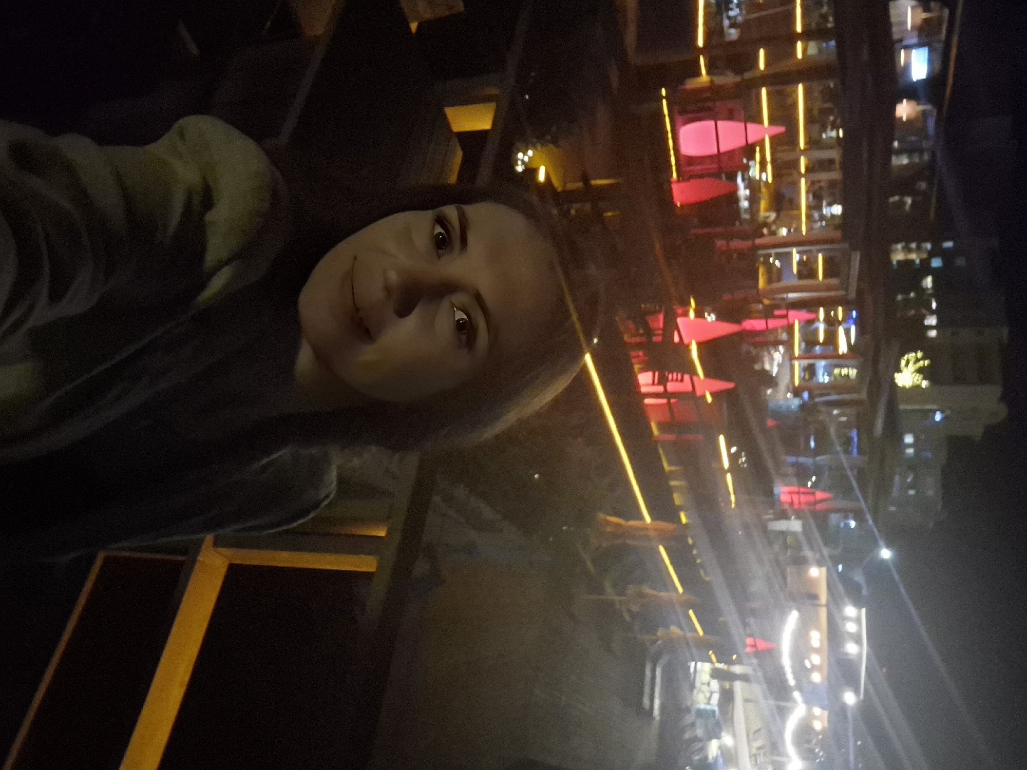 A selfie is taken at night by a young woman with brown hair and brown eyes, wearing a gray jacket. She is smiling and looking at the camera. The background is a nighttime scene of a restaurant or bar with red and yellow lights. Buildings with illuminated windows are visible in the distance. The scene appears to be outdoors, possibly on a patio or balcony. The location is Votkinsk, Russia.
