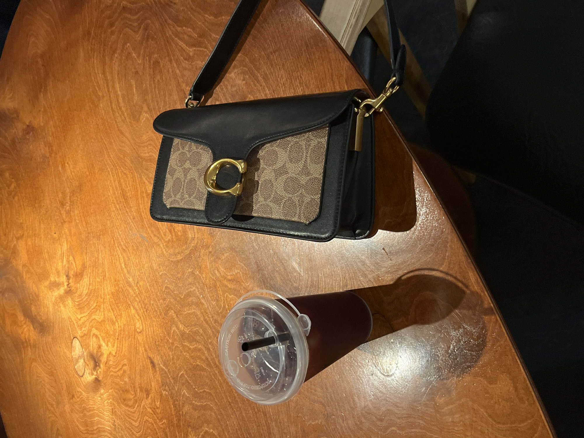 The scene is a close-up, high-angle shot featuring a wooden table. On the table sits a Coach handbag with a black leather flap and tan monogram canvas. A disposable coffee cup with a clear plastic lid and straw is positioned near the bag. The background shows a dark-colored surface, likely part of a chair. The location is indeterminate.