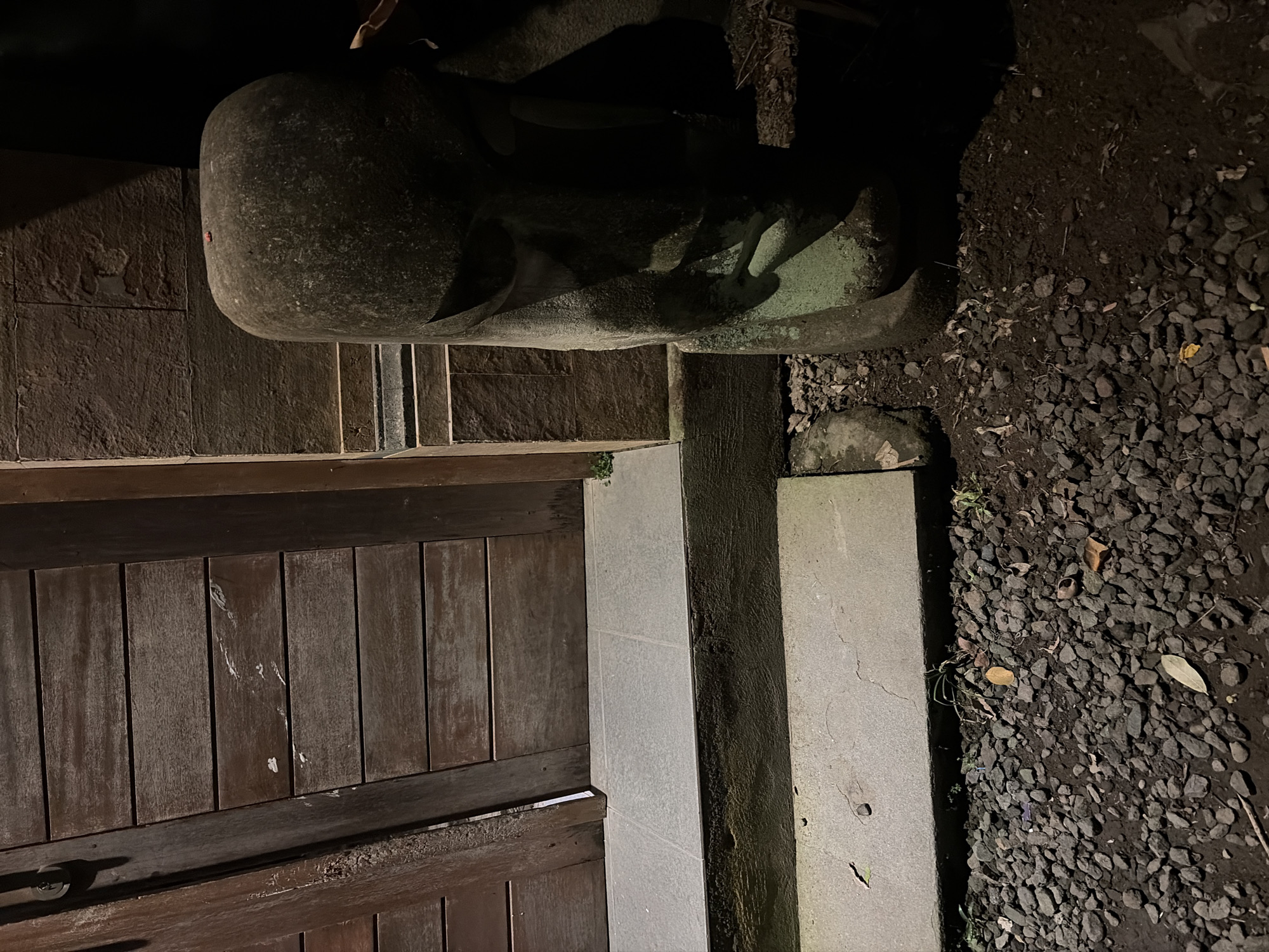 A stone statue, resembling a Moai head, stands to the right of a wooden door. The statue is weathered and has a green patina in some areas. It is positioned on dark soil with scattered small stones and fallen leaves. To the left of the statue is a tiled step leading up to the wooden door, which appears to be made of horizontal planks. The area is dimly lit, suggesting it is nighttime. The scene is located in North Kuta, Indonesia.
