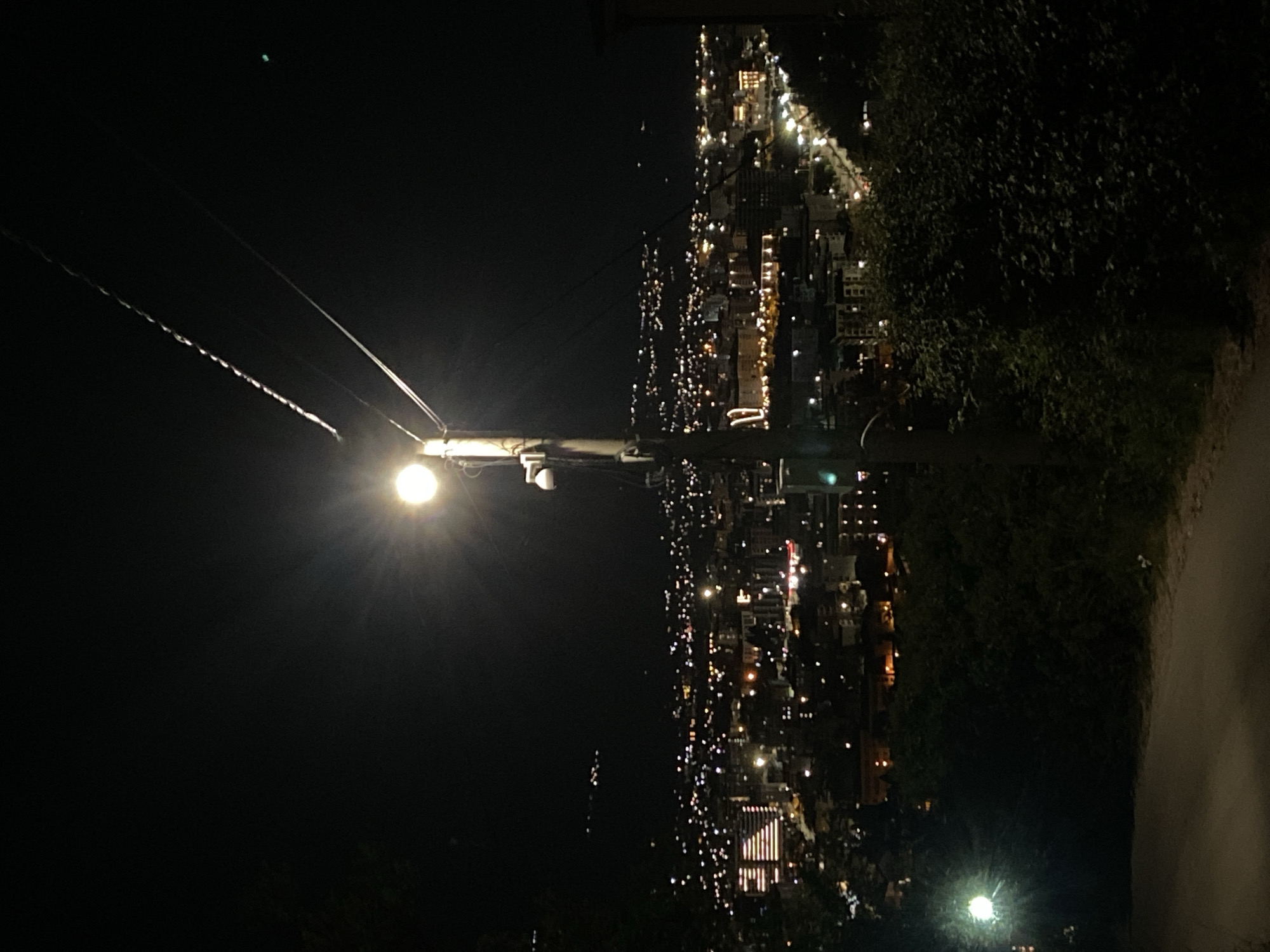 The image depicts a night view of a city, possibly Šušanj, Montenegro, based on the location provided. A street lamp is prominently positioned in the foreground on a utility pole. To the right of the pole, a dark, vegetated area leads to a lighted walkway. In the background, the city lights extend towards the horizon. A few powerlines stretch toward the sky.