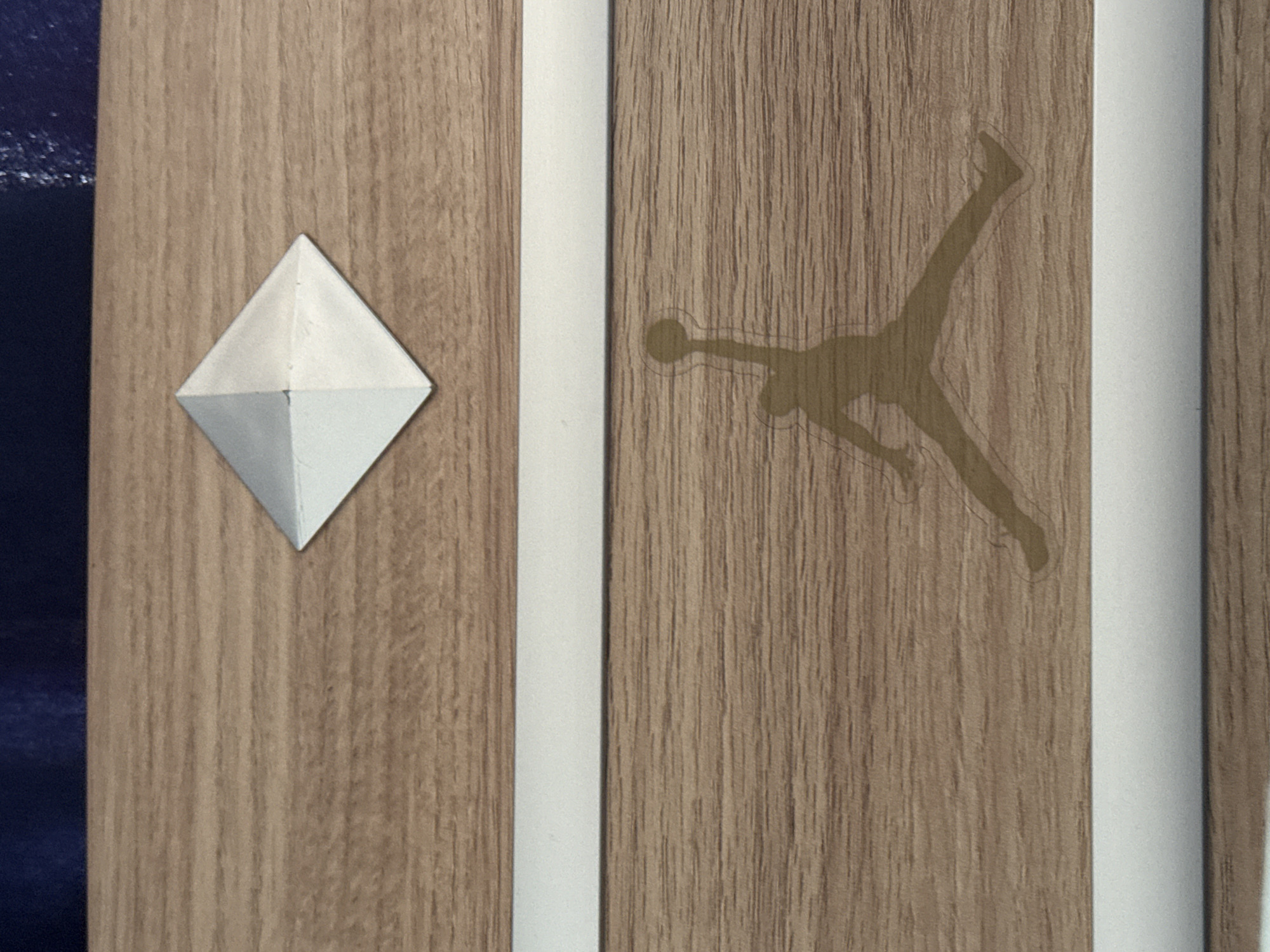 The image shows a close-up of a wooden surface with a silhouette of a basketball player mid-jump, commonly known as the Jumpman logo, and a silver diamond-shaped emblem. The surface appears to be a piece of furniture or packaging, with horizontal wood grain patterns. Two white horizontal stripes are visible, one above and one below the Jumpman logo. The Jumpman logo is positioned in the lower half of the image, and the diamond emblem is in the upper half. The background above the wooden surface is a dark blue, textured material.