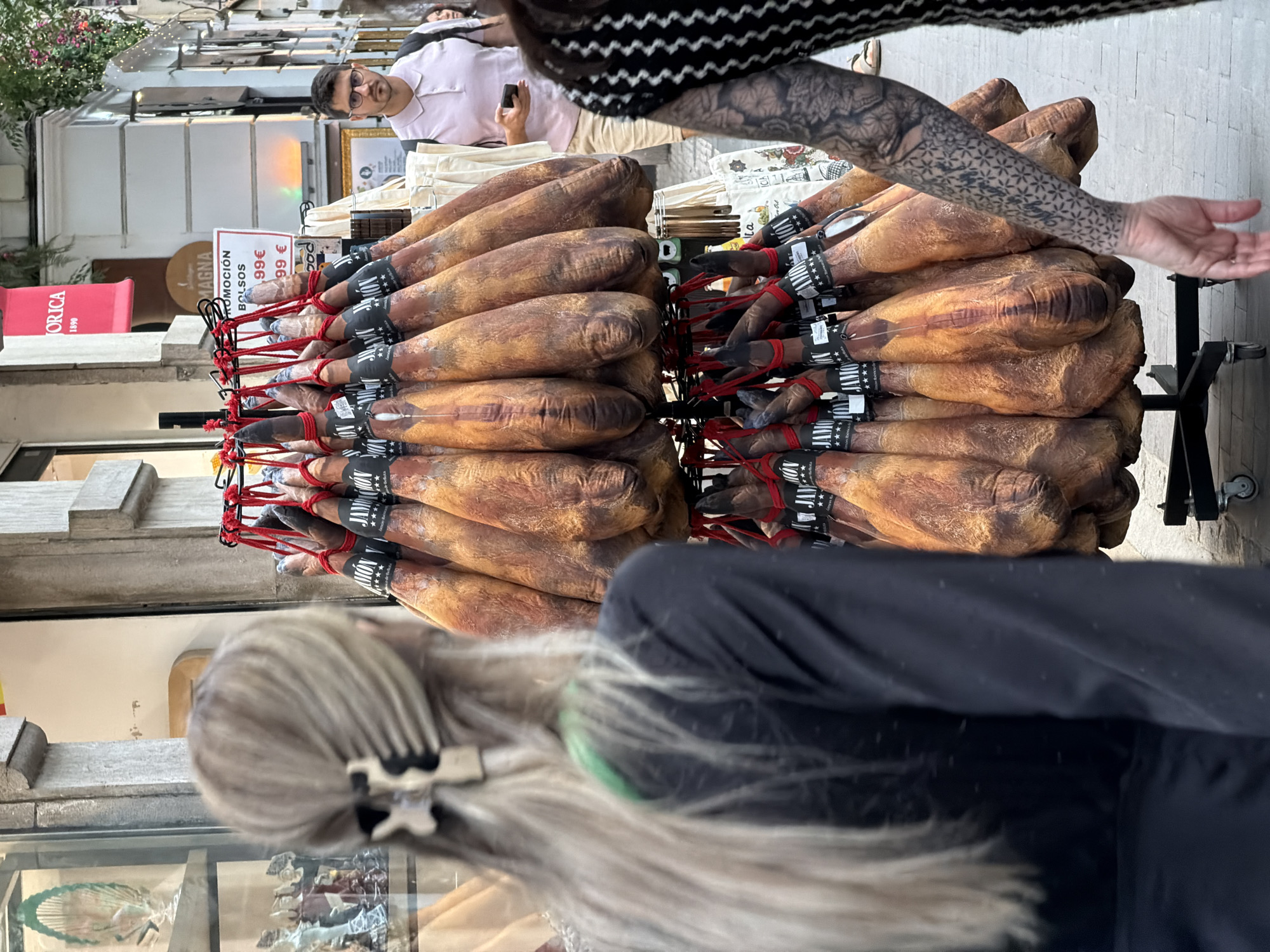 In an outdoor setting, a display of cured hams is presented, with some hanging from a metal rack. A woman with blonde hair pulled back by a clip is visible from behind, her left side facing the camera. To her right, a man with dark hair and glasses stands in a light pink polo shirt and tan shorts, holding a mobile phone. He is looking towards the camera. Another person's tattooed arm is visible in the foreground on the right. In the background, a red sign with white lettering is displayed. The environment appears to be a street or market area, with building facades and signage suggesting a commercial district.