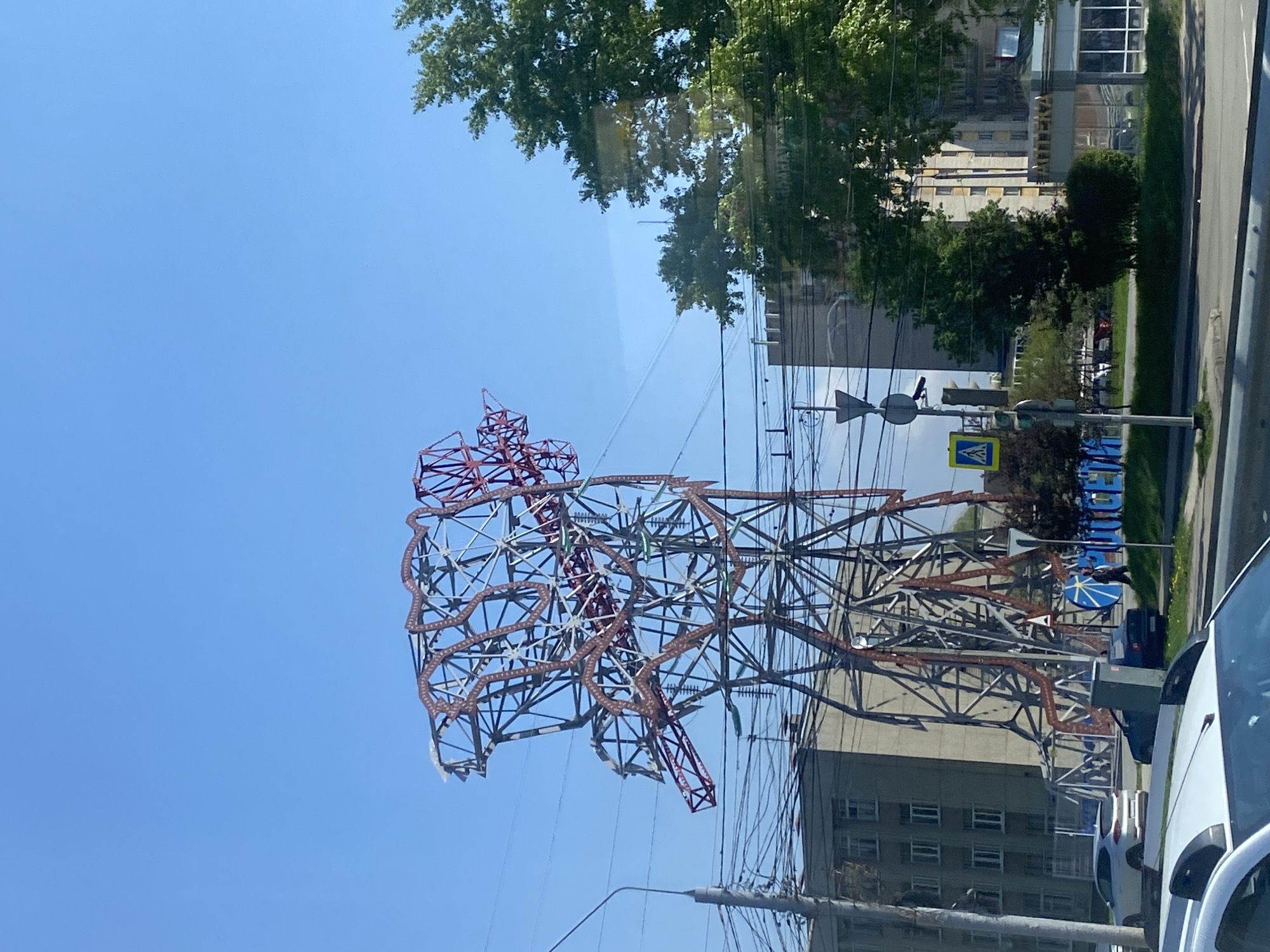 The image shows a distinctive, skeletal metal sculpture resembling the shape of a bear, standing in Rusanovo, Russia. The sculpture is constructed from intersecting metal beams, with red highlights outlining the bear's form. Overhead power lines cross the view in front of the bear sculpture. Adjacent to the sculpture are buildings and a road, including signs, a green space and at least 2 white cars. The sky is a clear blue.