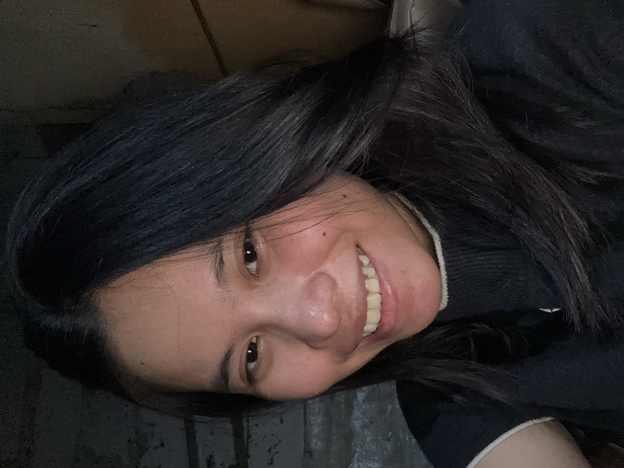 This is a selfie of a young woman with long, dark hair, smiling at the camera. She is wearing a dark, high-necked sweater with white trim on the collar and sleeves. The background is dimly lit and appears to be an indoor environment, possibly a room with textured walls. The overall lighting is soft, highlighting her face. No specific location within East Jakarta, Indonesia can be determined from the image.
