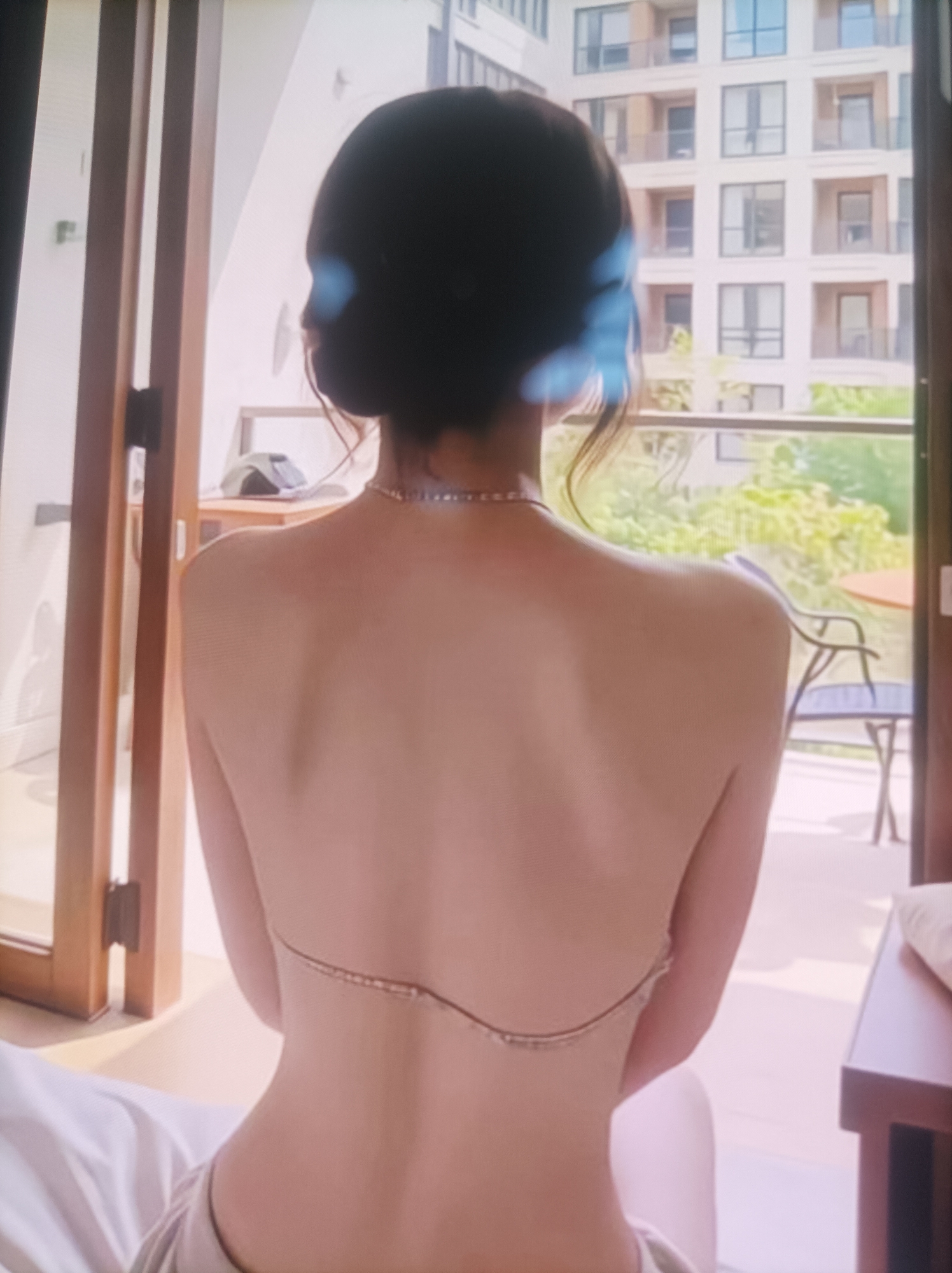 A woman with dark hair, seen from behind, is sitting on a bed, facing a balcony. She is wearing a top that exposes her back, which is adorned with a delicate chain. Through the open balcony doors, a building with multiple windows and a balcony with outdoor furniture can be seen, along with greenery. The room is lit by natural light, creating a bright and airy atmosphere.