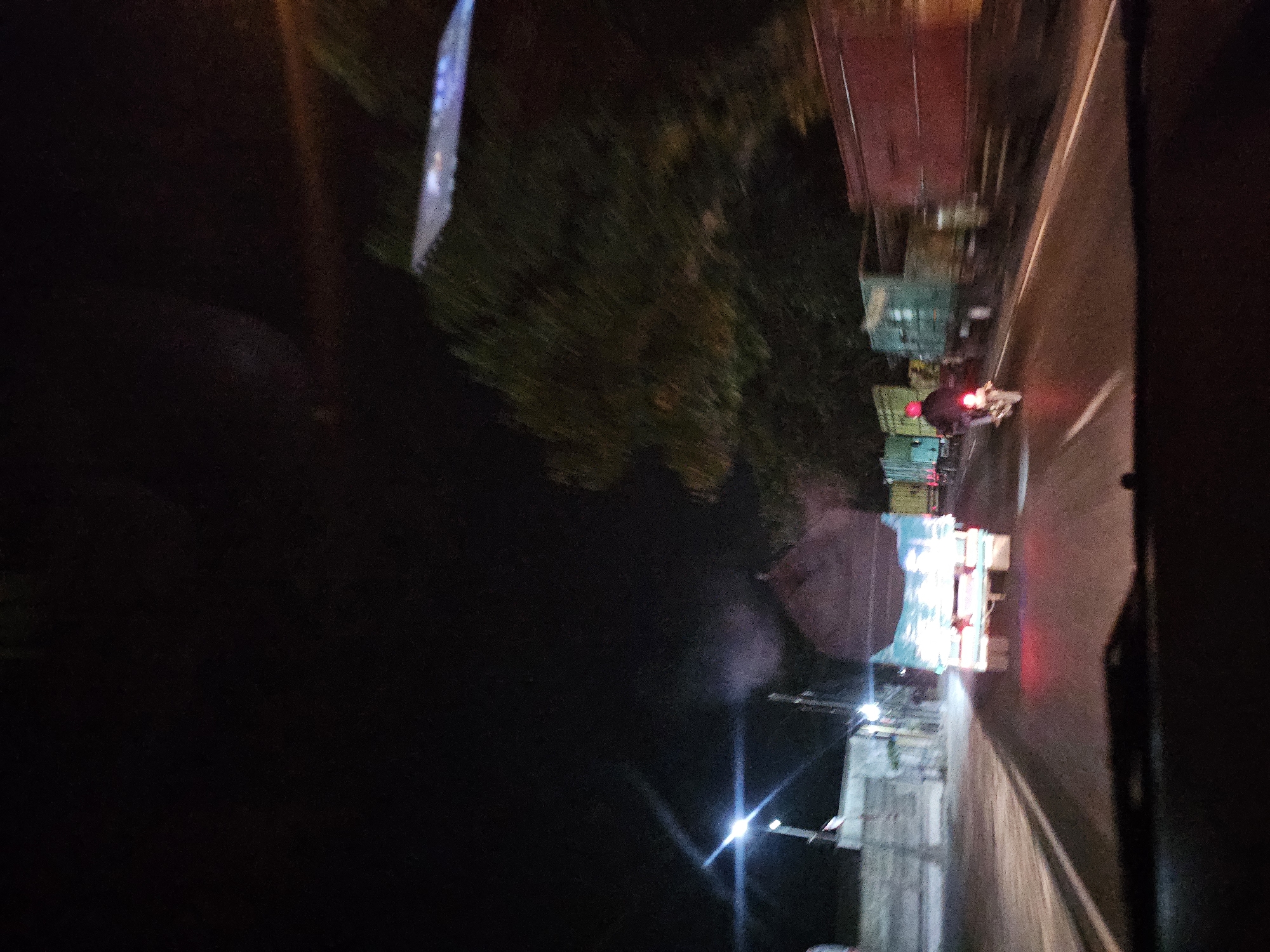 The image depicts a nighttime street scene in Waruwetan, Indonesia. The view is from the perspective of a vehicle, looking forward. The road is paved and has white lane markings. Several large trucks are visible, some carrying cylindrical loads, and one appears to be a flatbed truck. One truck in the foreground has its taillights illuminated red, indicating it is stopped or moving slowly. To the right of this truck, a motorcycle with a rider wearing a red helmet is passing. The rider's taillight is also red. Trees line the right side of the road, and foliage is present. On the left side of the road, there are buildings and streetlights, one of which is emitting a bright white light. The overall atmosphere is dark, characteristic of nighttime.