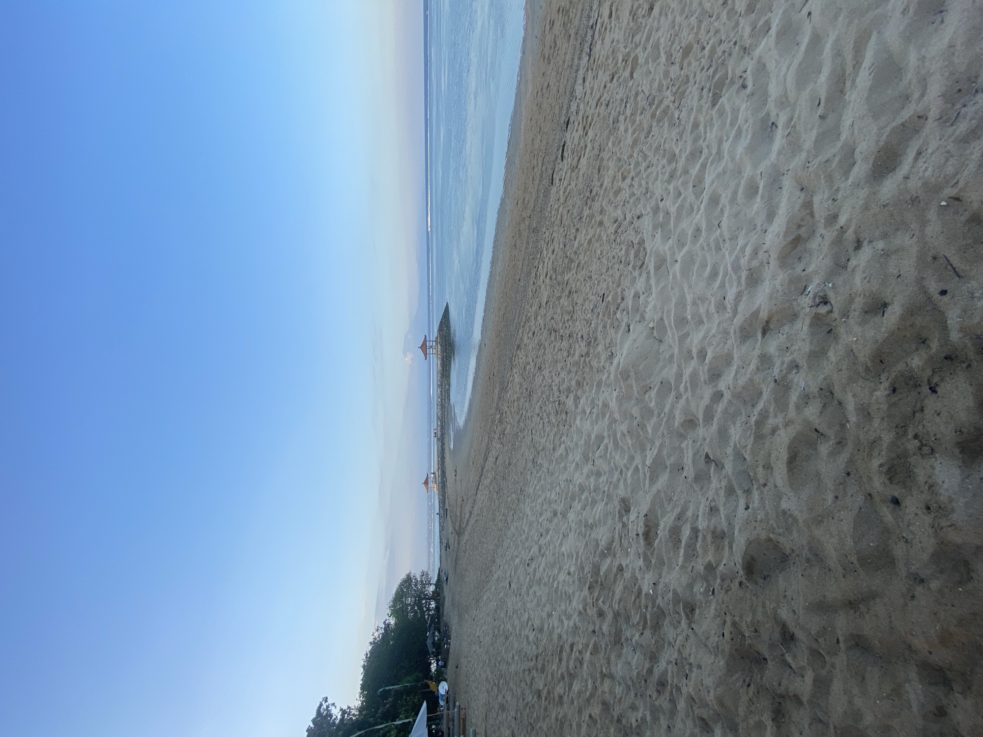 This image depicts a serene beach scene at sunrise or sunset, likely in Denpasar, Indonesia, given the presence of a traditional Balinese gazebo or bale bengong. The foreground is dominated by a wide expanse of light-colored sand, textured with ripples and small debris, indicating the ebb and flow of the tide. In the mid-ground, the calm, blue ocean stretches to the horizon, meeting a clear, pale blue sky. Along the waterline, a man-made breakwater or jetty extends into the sea, topped with a small, red-roofed gazebo. Further down the beach, on the left side, are a few scattered beach umbrellas, some tables, and what appear to be lounge chairs, suggesting a developed tourist area. A cluster of green trees and foliage marks the transition from the beach to the land. The overall atmosphere is peaceful and tranquil, with no people visible in the frame, highlighting the natural beauty of the location.