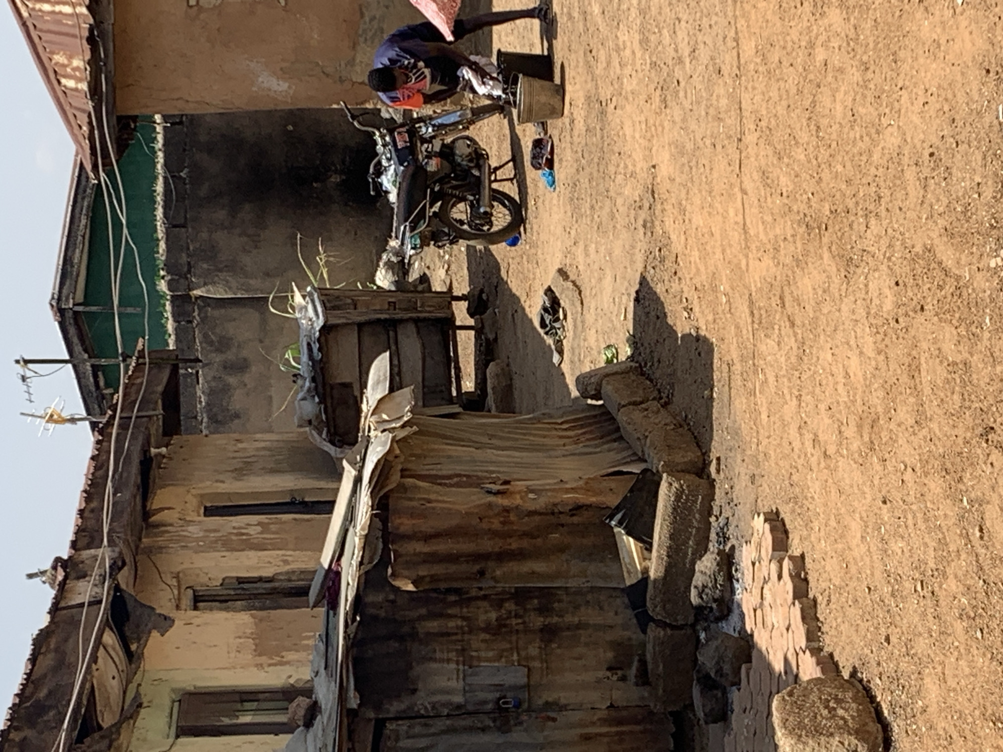 In Rafin Par, Nigeria, a narrow, sunlit alleyway separates dilapidated buildings constructed of aged brick and corrugated metal. A person washes clothes on a motorcycle parked in the alley, using a bucket and other containers. The ground is dirt with areas of brickwork. Overhead, an antenna is visible above the crumbling rooftops.