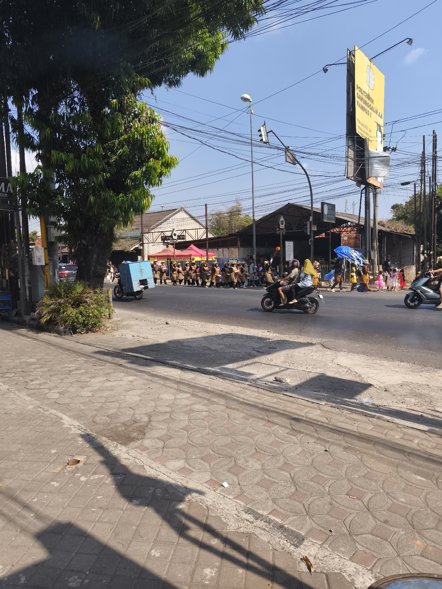 In Kutoarjo, Indonesia, a street scene unfolds under a clear blue sky. A procession or parade is in progress, with a group of individuals in brown and tan uniforms marching on the right side of the asphalt road. Some are carrying what appear to be instruments or flags. Further down the road, children in colorful costumes are visible, some being guided by adults.
Several vehicles are present on the road. A scooter with two occupants, one wearing a yellow headscarf, is moving past the marching group. Another scooter, ridden by a single person wearing a helmet, is further ahead on the right. In the foreground on the left, a blue box is attached to the back of a scooter being ridden by a person wearing a helmet. Black electrical wires are strung across the sky, and traffic lights are visible.
Buildings line the street, including one with a brown roof in the background. Several red and pink tents are set up along the side of the road, possibly for vendors or spectators. A tall yellow billboard with text in Indonesian is prominent on the right side of the image. The sidewalk in the foreground is paved with patterned gray and red tiles, and shadows cast by trees and possibly overhead structures fall across it. Some greenery, including a large tree and bushes, is visible on the left side of the frame.