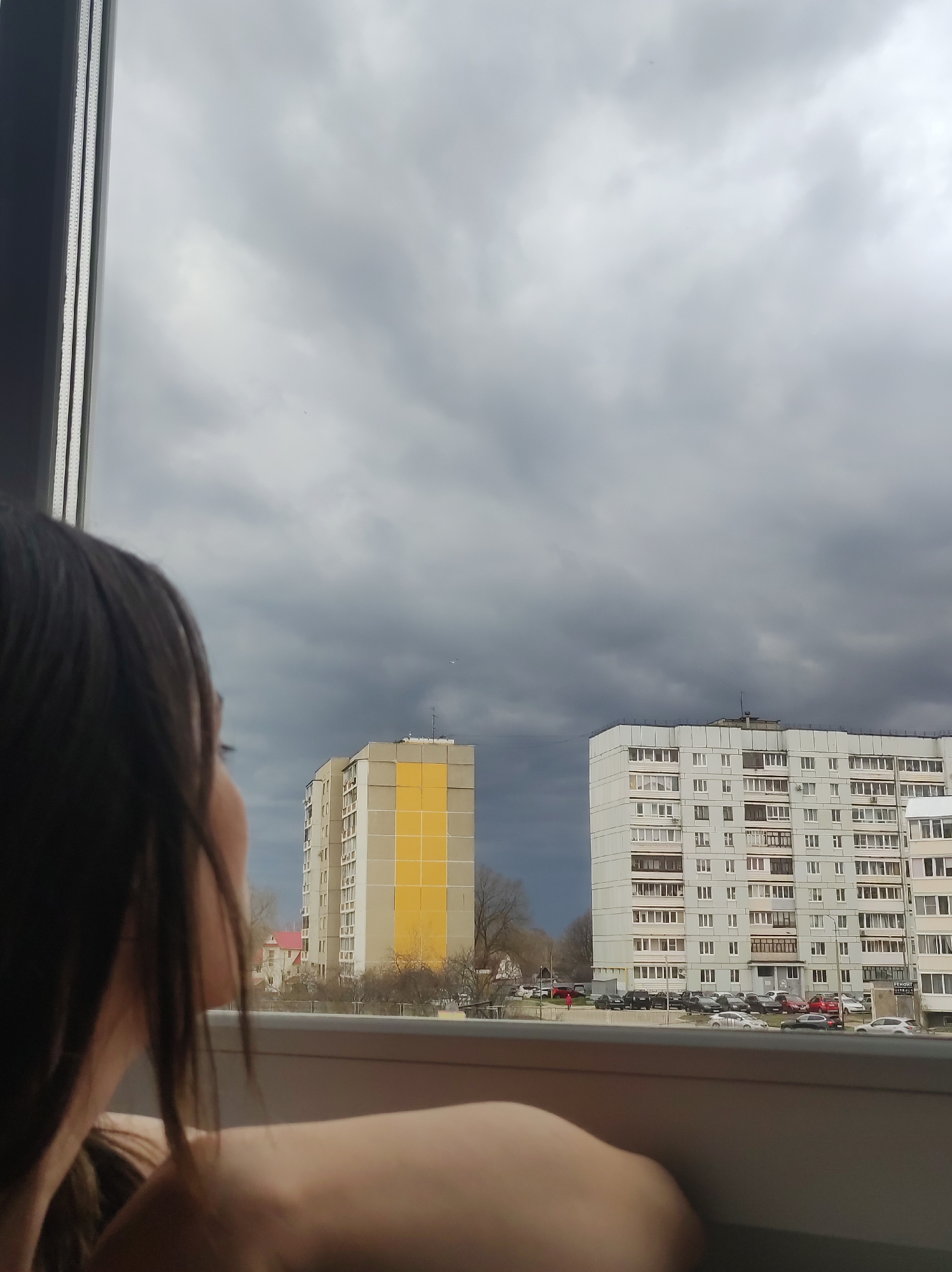 The image is an interior shot taken from what appears to be a window. A person is partially visible in the foreground, with their back and head taking up the left side of the frame. They are looking out the window. Outside, there are two apartment buildings of typical Soviet-era construction. The building on the left is distinguished by a large yellow section on its facade. Numerous parked cars are visible in front of the buildings. The sky is overcast with dark, ominous clouds. Given the architectural style of the buildings, this scene is likely in a post-Soviet city, possibly the city of Chaikovsky in Russia.