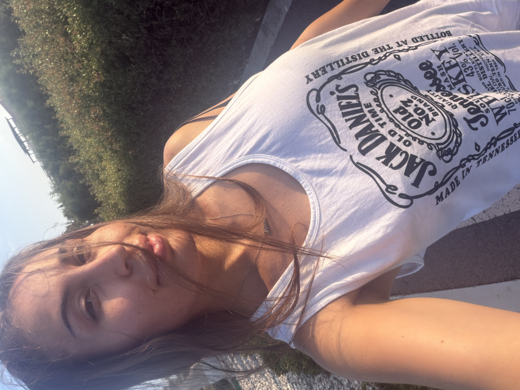 A young woman is taking a selfie outdoors, with a blurred background of vegetation and a clear sky. She is wearing a white tank top with a Jack Daniel's logo. Her hair is brown and falls over her face, obscuring some of her features. The lighting suggests it is either sunrise or sunset. The location is in Meneou, Cyprus.