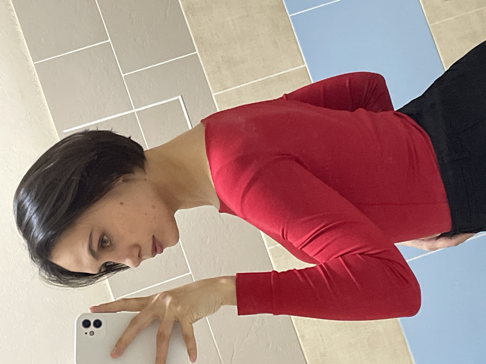 A person is taking a selfie with a white smartphone in what appears to be a bathroom. The person has short, dark hair and is wearing a red long-sleeved shirt and dark pants. The background shows beige and light blue tiles. There is no indication of this being in Izhevsk, Russia.