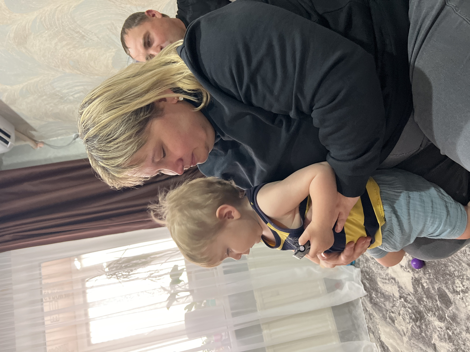 The image shows a family scene, likely indoors. A woman with blonde hair, wearing a black hoodie, is holding a young child in her arms. The child, who also has blonde hair, is wearing a yellow and navy tank top with light blue shorts, and is holding a toy car. A man is partially visible behind the woman, his face showing above her head. The setting appears to be a home interior, with a window covered with sheer curtains in the background. There is also what seems to be a piece of furniture draped with a brown cloth to the left of the window. A purple ball is lying on the floor. Noviy, Russia, is not specifically detectable in this image, as interior settings generally lack location-specific indicators unless there is distinctive architecture or signs of the locale.