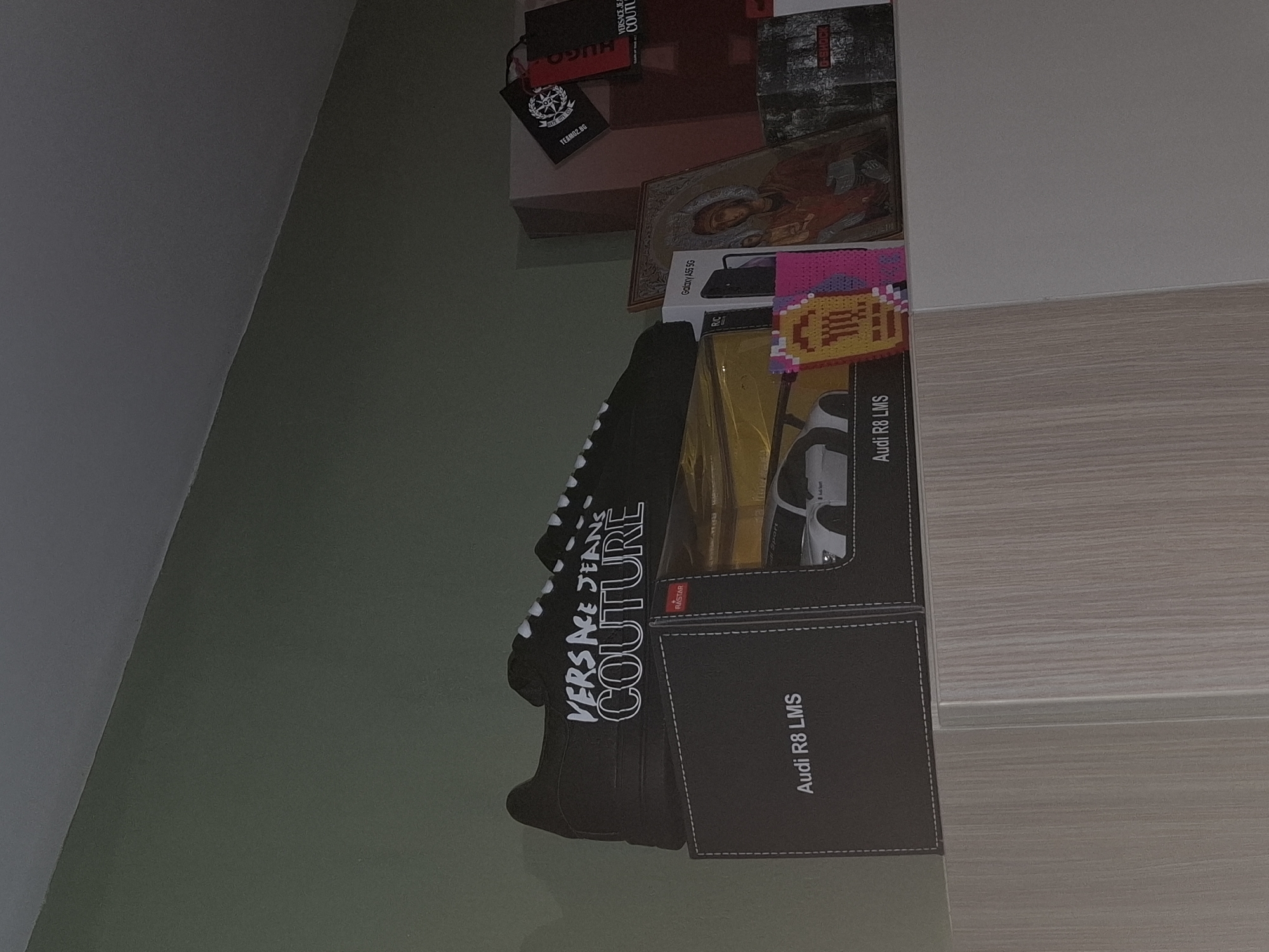 The image shows a collection of boxes and other items stacked on a surface. There are clothing brand boxes from Hugo and Versace, along with a box for a model of an Audi R8 LMS sports car. The boxes appear to be stacked inside a closet or storage space, as the image also reveals a wall corner and a wooden surface with horizontal grain. The environment around the items appears to be dimly lit. There is no information to suggest this image was taken in Veliki Preslav, Bulgaria.