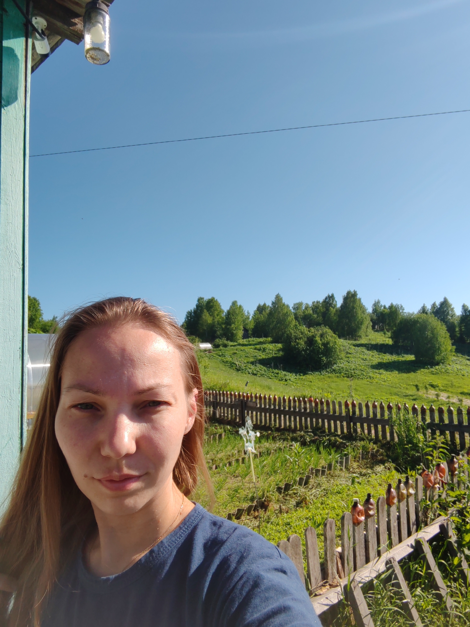 A self-portrait captures a woman in the foreground against a rural landscape in Чухлэм, Russia, under a clear blue sky. The woman, with long, light brown hair and a fair complexion, wears a dark blue short-sleeved t-shirt and a thin gold chain, looking directly at the camera with a neutral expression. Her face is brightly illuminated by direct sunlight.
To the viewer's left, a vertical wooden beam, painted a faded teal and showing signs of peeling paint, marks the edge of a structure. Above this beam and slightly to the right, an outdoor light fixture with a clear cylindrical glass casing and visible debris inside is attached to a wooden eave. Next to it, a white electrical component is affixed. A thin black wire extends horizontally across the upper portion of the clear blue sky.
The midground features a rustic wooden picket fence, with numerous plastic bottles, some brown/orange and others white, placed atop many of its pickets. Beyond the fence, a gently sloping hillside is covered in vibrant green grass and dense clusters of deciduous trees with full, bright green foliage. A translucent greenhouse structure is partially visible behind the woman's head, and another smaller white structure appears further up the hill. The ground in the immediate foreground consists of overgrown grass and weeds. The scene is brightly lit by direct sunlight, casting distinct shadows.