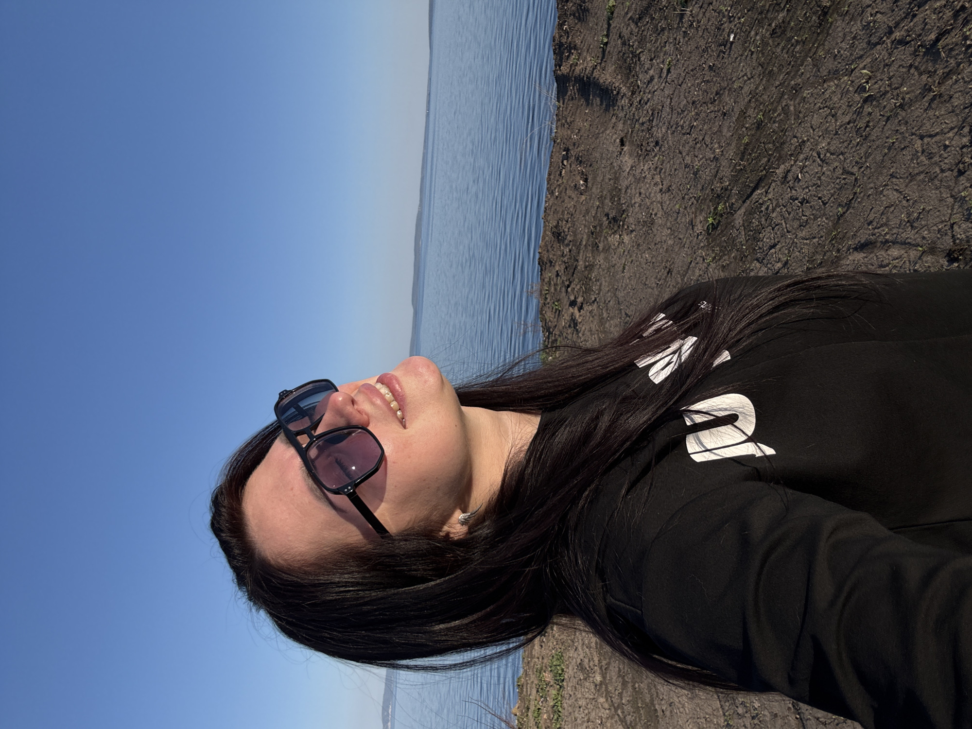 A woman with dark hair and sunglasses is looking upwards in a selfie taken outdoors. She is wearing a black top with white lettering. The background shows a blue sky, a body of water, and a patch of dry land. The location is Unknown, Unknown.