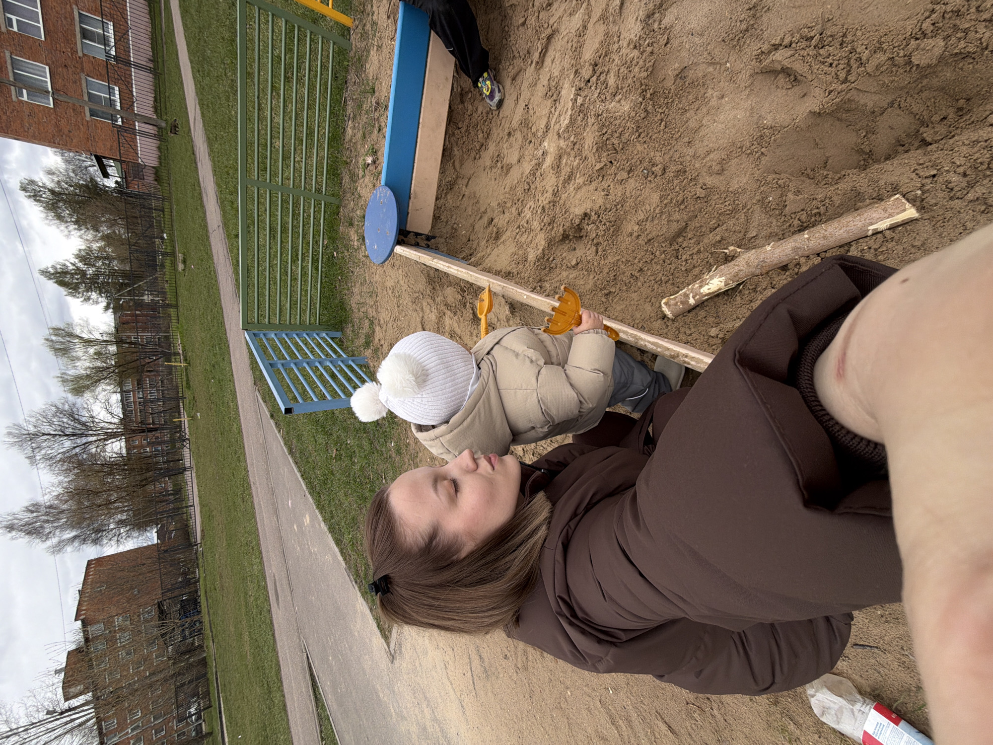 The image depicts an outdoor scene in Reshetnikovo, Russia. A woman in a brown jacket is lying on the ground in a sandbox. Nearby, a child in a beige jacket and white hat is holding a toy shovel. In the background, playground equipment including a see-saw and a small blue gate are visible. Beyond the playground, residential buildings and trees are seen. The sky appears overcast.