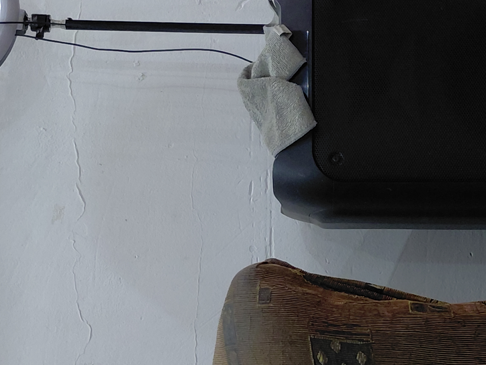 An indoor scene, potentially in Santa Ana, Venezuela, captured from a slightly elevated angle, features a white, textured wall as the dominant background. The wall shows signs of unevenness and several subtle cracks or lines running vertically and horizontally.
In the upper left quadrant, a black horizontal bar or rod is mounted to the wall, secured by dark fixtures. A thin black wire runs horizontally beneath this bar. Draped over the black bar and resting against a larger black object in the upper right is a light-colored, possibly grey or beige, folded cloth with a visible textured surface, resembling a small towel or rag.
The large black object, occupying the upper right and extending downwards, appears to be an electronic device, perhaps a speaker or part of a monitor. It has a finely textured, almost mesh-like surface and a visible screw or fastener near its bottom left edge.
In the lower right portion of the image, a segment of a brown, highly textured object is visible. This object has distinct vertical ridges and some darker, possibly decorative, square-like patterns. Its upper edge appears rounded, suggesting it could be part of a woven basket, a piece of furniture, or an artifact made of wood or natural fibers.
There is no activity taking place, and no people are visible. The lighting suggests an indoor setting but offers no specific cues about the time of day or external weather. There is no visible text or explicit location information within the image itself.