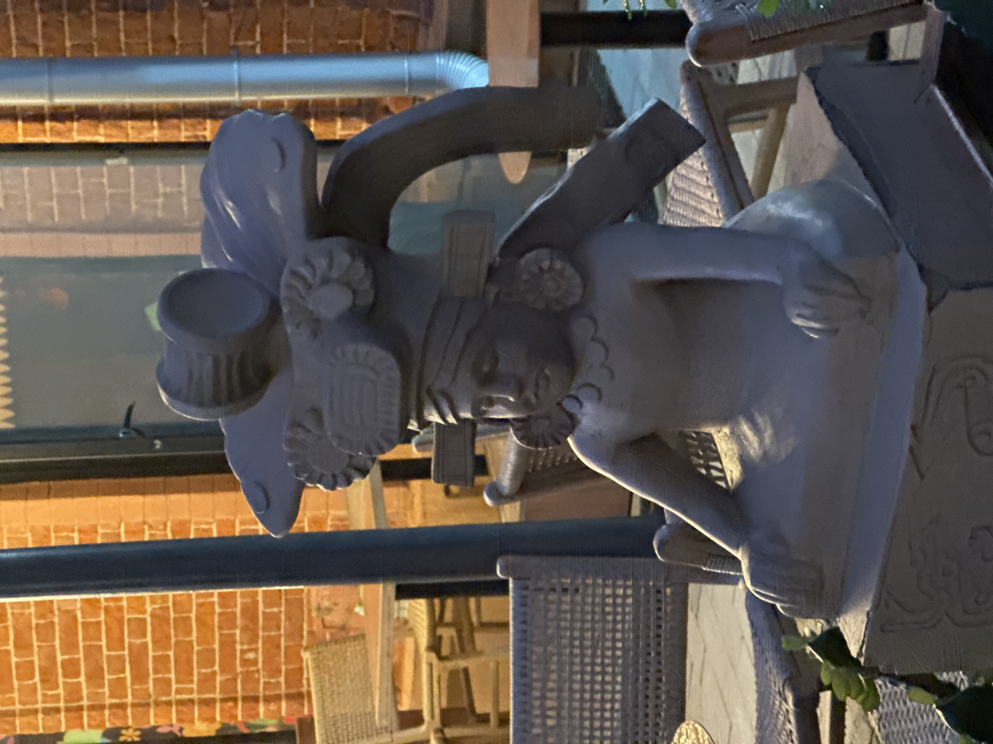 A statue of a Mayan figure is in a courtyard area. The figure is seated with its hands on its lap. It wears a headdress that resembles a headdress with two circular ear ornaments. The statue is made of a light-colored material and is positioned on a square base with carved designs. The background shows a brick wall, outdoor seating with wicker chairs and tables, and a metal pipe. The scene appears to be in an outdoor dining or seating area, possibly at a restaurant or cafe. There are no people visible in the image. The statue is the central focus, with the surrounding environment providing context. The lighting suggests it may be evening or dusk.