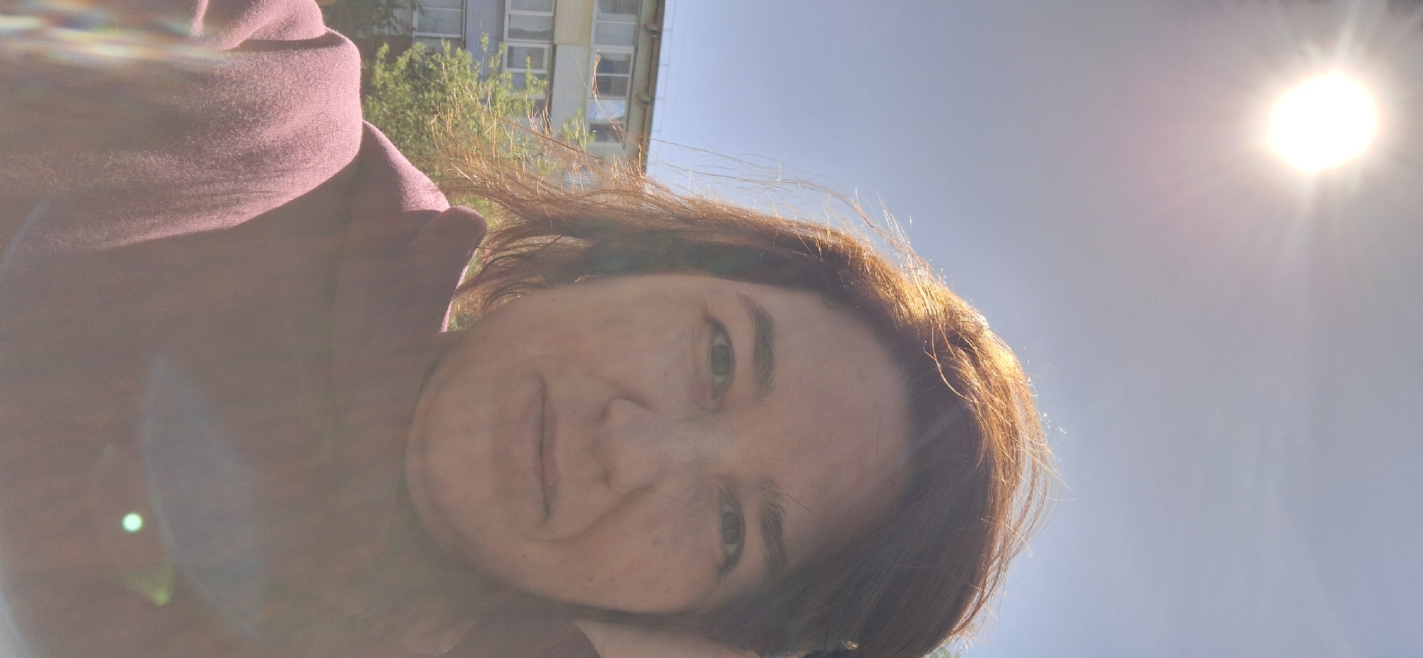 A selfie of a woman with light brown hair, wearing a maroon top, is oriented diagonally against a bright, sunny sky. The intense sunlight washes out some details of her face. In the background, a multi-story building with multiple windows is partially visible. The photo may have been taken outdoors in the city of Chaikovsky, Russia. The focus is on the woman’s face and upper body, with the sun as a prominent feature in the upper right.