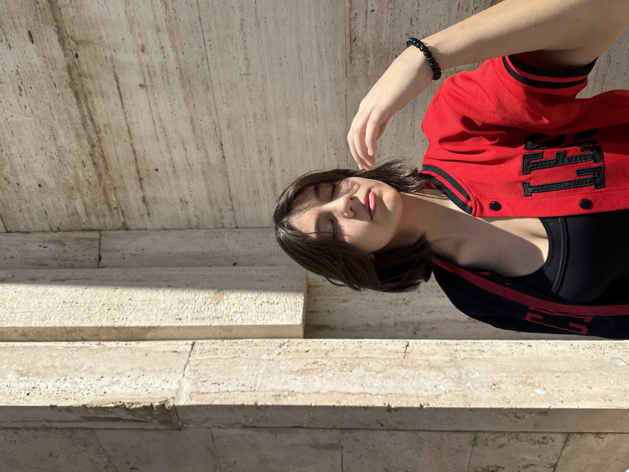 A young woman lies on a set of stone steps. She has short brown hair and is wearing a red and black jacket over a black top. Her eyes are closed and her hand rests near her face. She wears a black beaded bracelet on her wrist. The steps are made of light-colored stone. The location is Yerevan, Armenia.