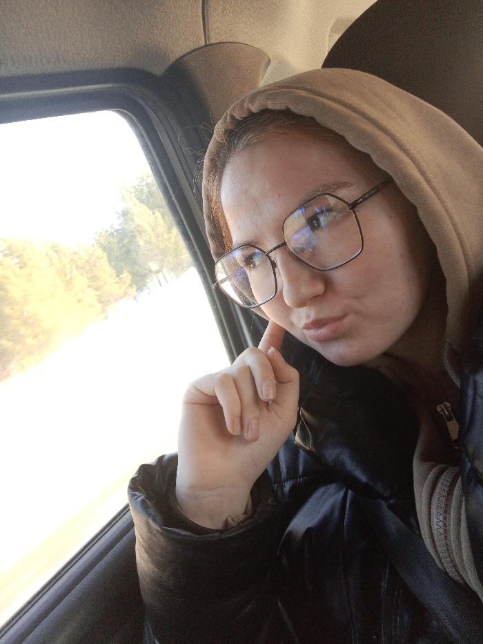A young adult female is depicted in a close-up, selfie-style photograph taken inside a moving vehicle in Krasnoyarsk, Russia. The individual, positioned off-center to the right, has fair skin, light brown hair partially concealed by a beige or tan hood, and wears dark-framed, rectangular eyeglasses that reflect ambient light. Her lips are slightly pursed, and her right hand, with a natural manicure, is raised with the index finger touching her right cheek. She is dressed in a dark, possibly black, quilted or padded jacket worn over a lighter, hooded garment. The vehicle's interior features a beige headliner and a dark grey window frame. Through the passenger window, a bright, overexposed outdoor scene is visible, characterized by blurred yellowish-green foliage and trees, indicating daytime and vehicle motion.