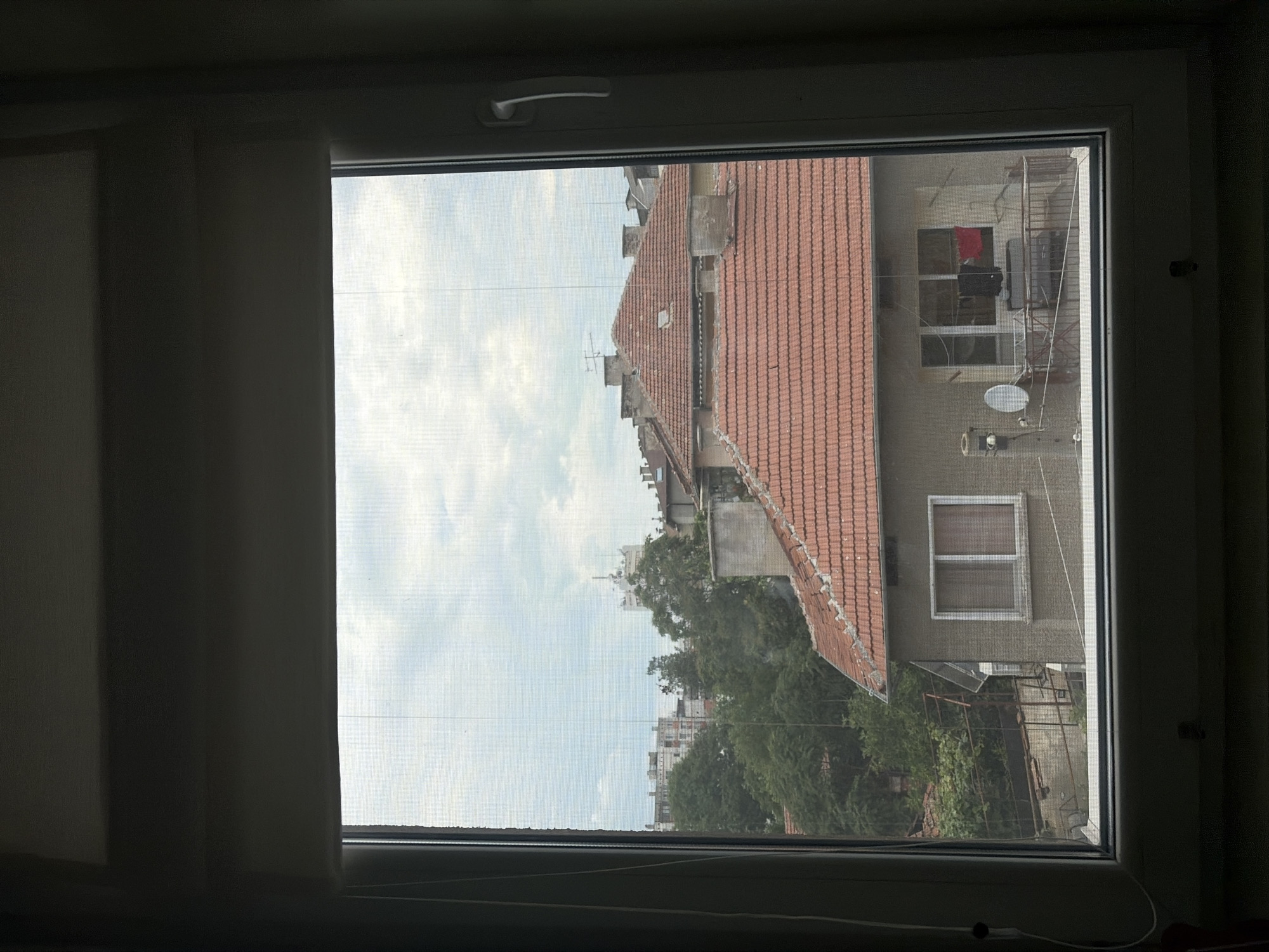 The media file shows a view through a window with a screen. The scene is outdoors, with a cloudy sky and buildings visible. The image is oriented vertically, with the sky taking up a significant portion of the left side and the buildings on the right.
The buildings appear to be residential, with terracotta tiled roofs and visible windows. One building has a red object, possibly clothing, hanging on a line outside a window. There are also trees and greenery visible in the background. A satellite dish is mounted on the side of one building. The window itself has a white frame and a handle. A sheer curtain is partially drawn on the left side of the window.
The lighting suggests it is daytime, likely overcast due to the clouds. There are no people clearly visible, but the presence of hanging clothes implies human activity. The setting appears to be an urban or semi-urban environment. No text is visible in the image.