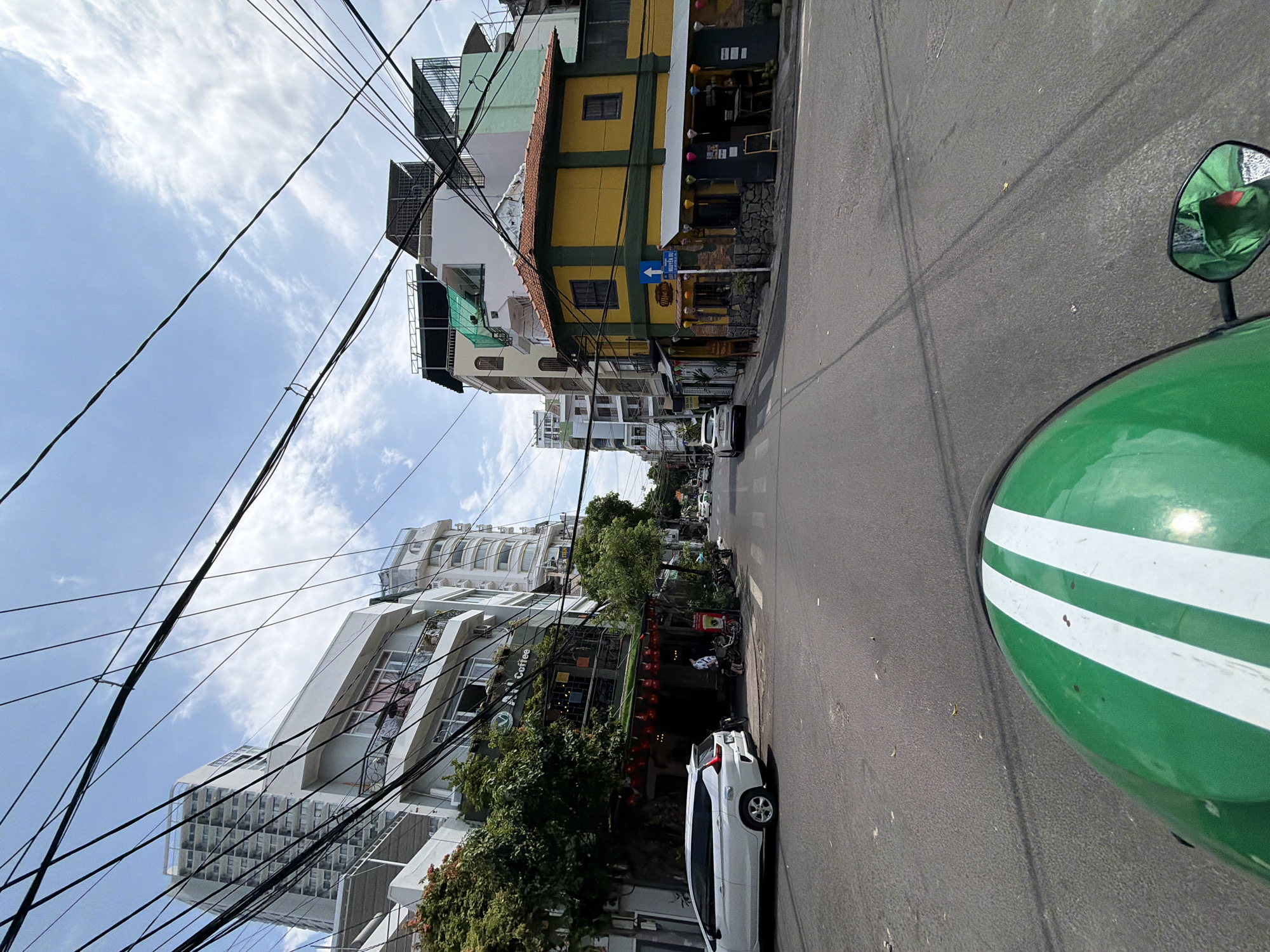 This image depicts a street scene in Nha Trang, Vietnam, likely taken from the perspective of a motorcycle rider. The foreground features the top of a green helmet with white stripes. The street is paved and appears to be a moderately busy intersection with several buildings lining both sides.
On the left, a white car is parked near a multi-story building with balconies and trees. A coffee shop with a green awning and hanging red lanterns is visible at street level. Further down the street on the left, taller, modern buildings with numerous windows can be seen.
On the right side of the street, a distinct yellow building with a green trim and a tiled roof stands out. It has a street sign indicating "NGUYỄN DU". There are tables and chairs set up outside what appears to be a restaurant or cafe. Further down the street, more buildings and some traffic are visible.
The sky is blue with scattered white clouds. A dense network of overhead electrical wires crisscrosses the entire scene, a common characteristic of urban Vietnamese infrastructure. The overall impression is a typical day in a bustling Vietnamese city, with a focus on the urban landscape and its infrastructure.